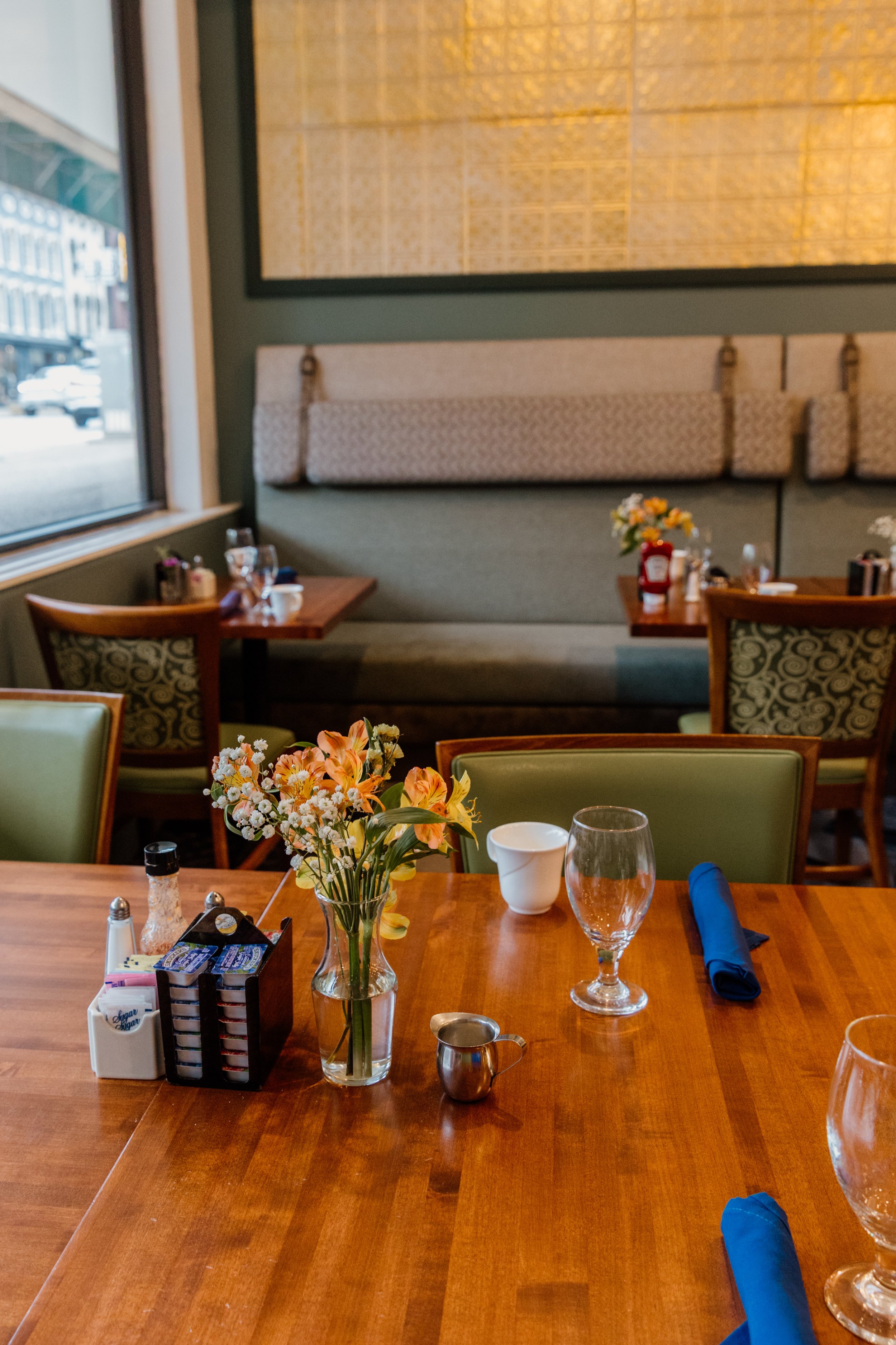 A table in a restaurant with a vase of flowers, wine glasses, a cup, a small pitcher, salt and pepper shakers, sugar packets, and blue napkins.