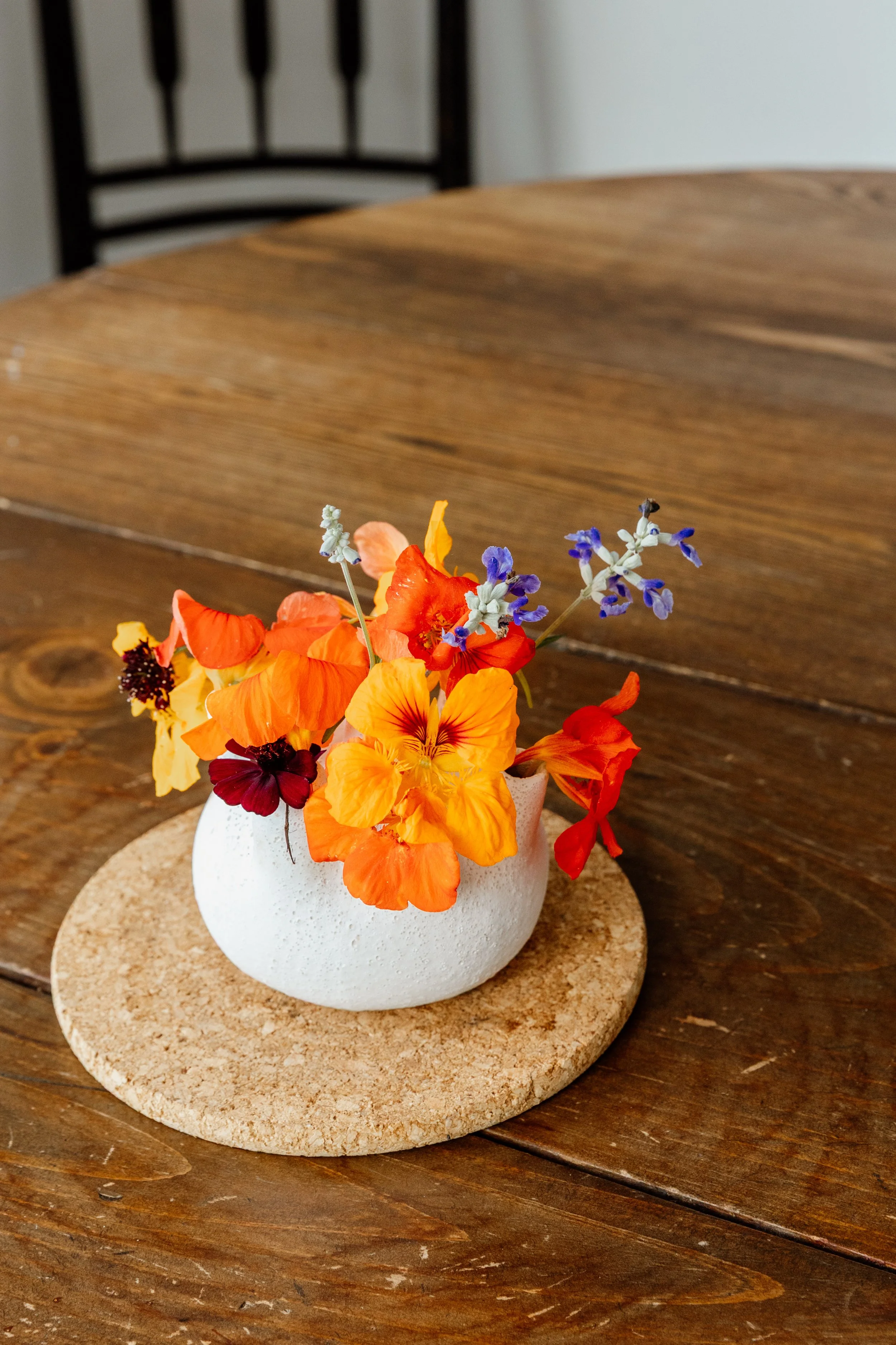 A small white ceramic vase with colorful flowers, including orange, yellow, red, and purple blooms, placed on a round cork coaster on a wooden table. A black chair is in the background.