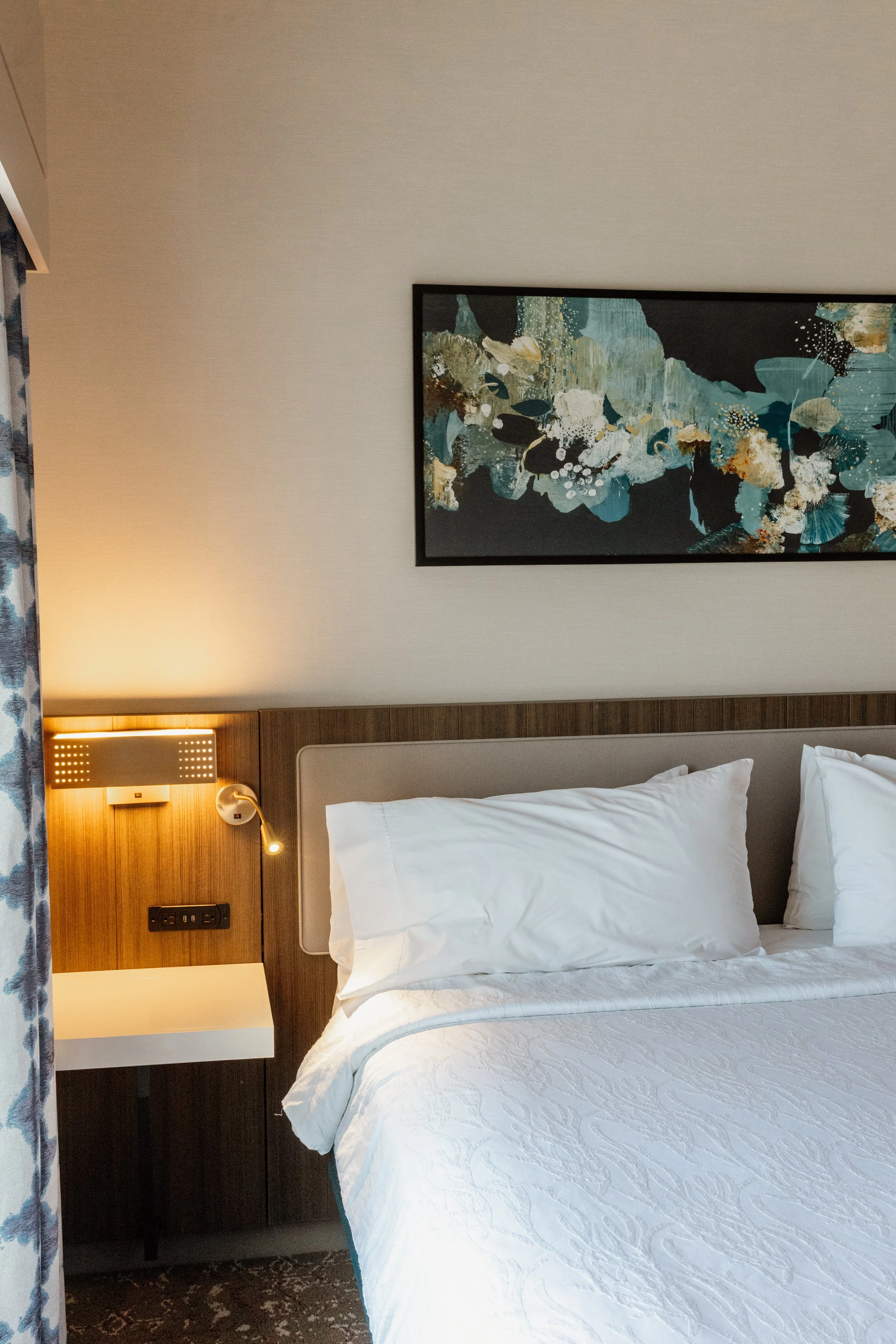 A neatly made hotel bed with white pillows and a quilt, a wall-mounted wooden headboard, a bedside lamp, a framed abstract floral painting above the bed, and a curtain on the left side of the image.