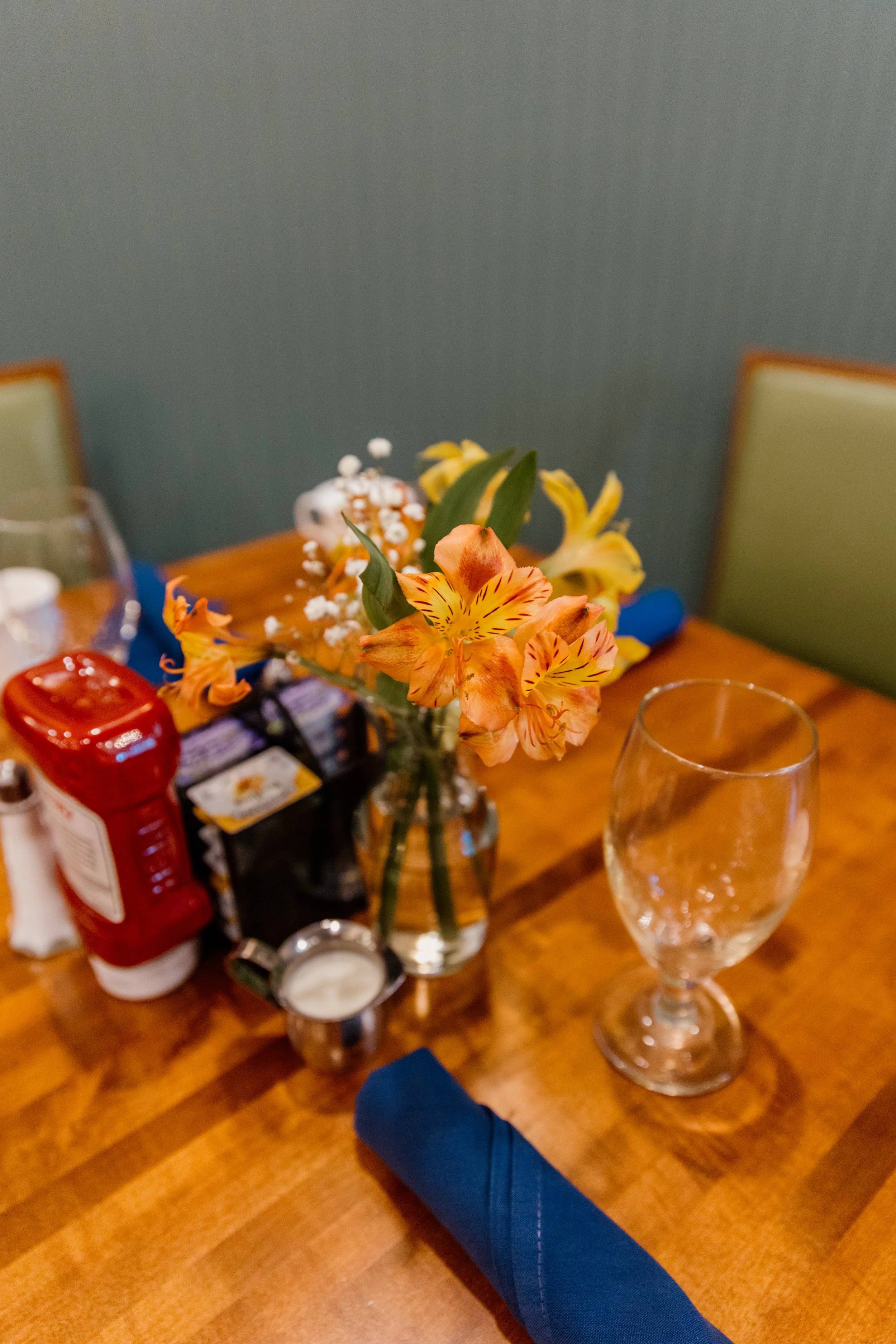 A wooden table with a small vase of orange and yellow flowers, a glass of water, blue napkin, ketchup bottle, and packets of sugar or sweetener, with green and beige chairs in the background.