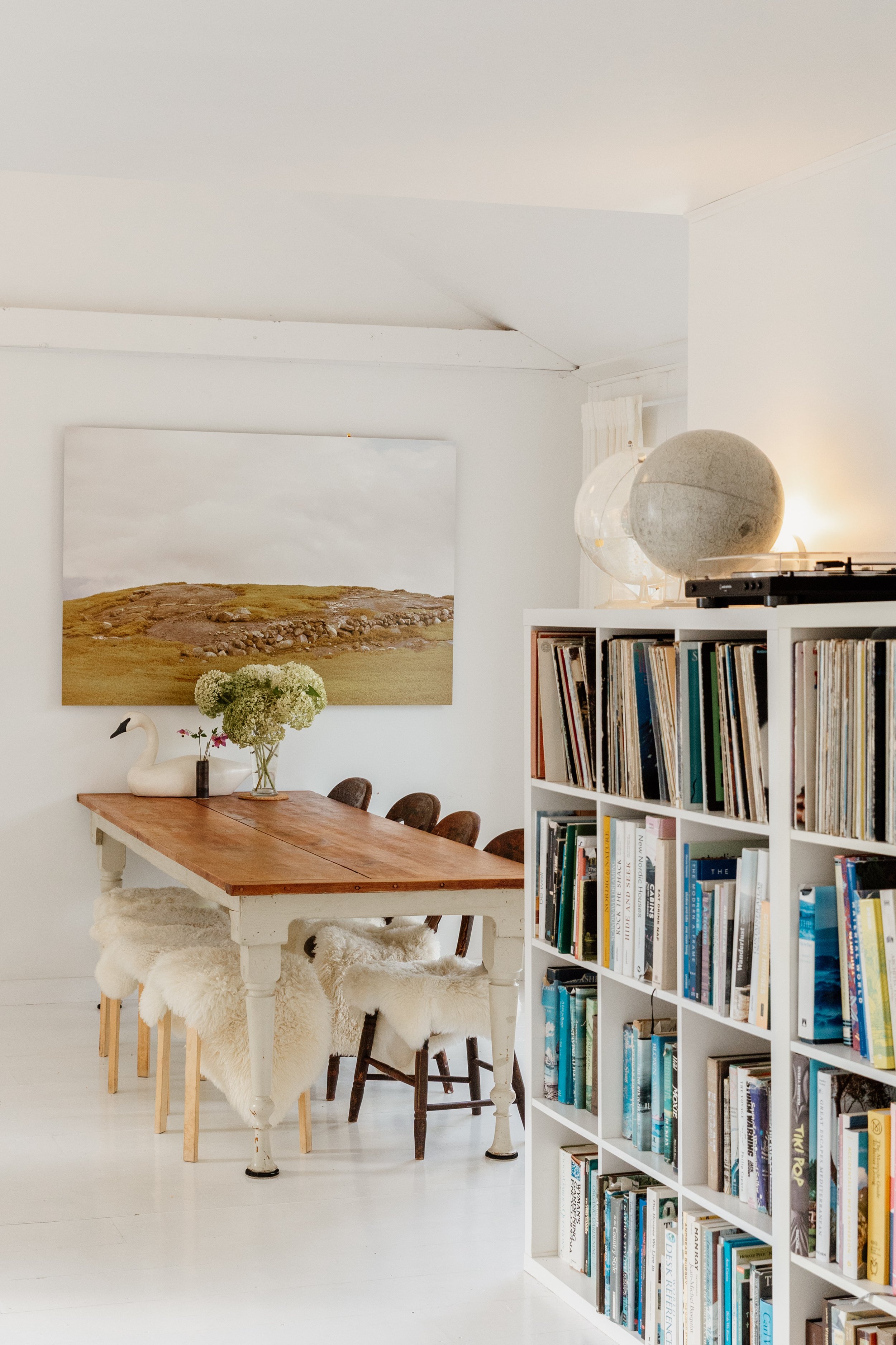 A cozy dining area with a wooden table, surrounded by chairs with fluffy white covers. A large landscape painting hangs on the white wall behind the table. On the right, there's a white bookshelf filled with books, with decorative globes and a turnta