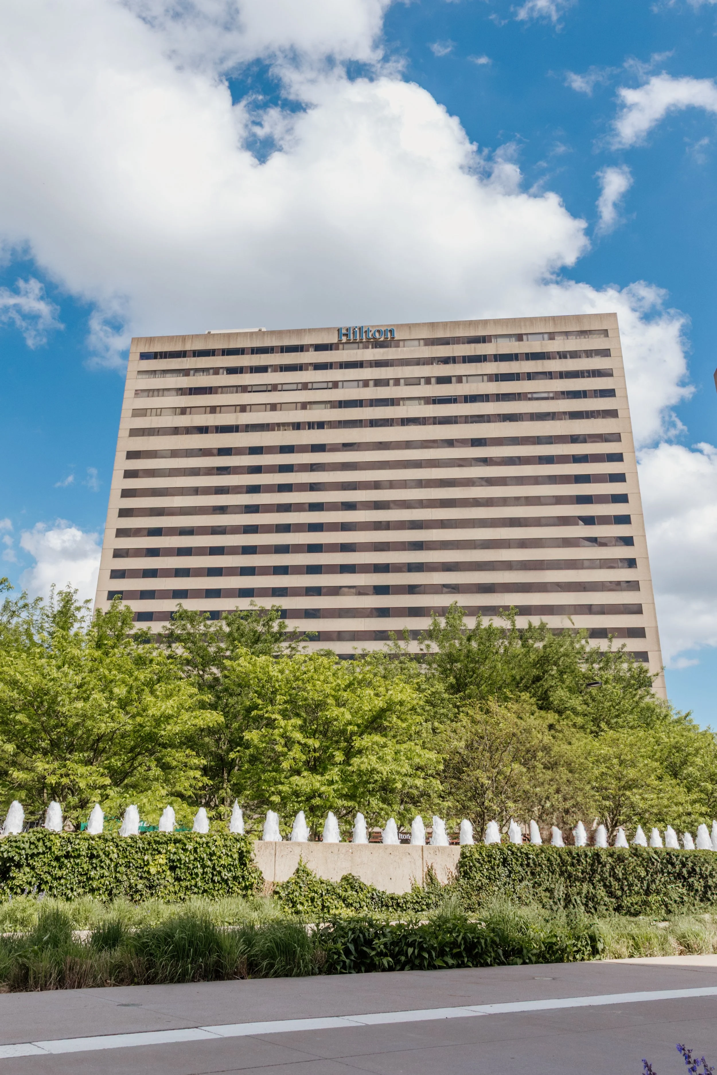 Tall Hilton hotel building with the name at the top, surrounded by green trees and a water fountain feature in front, under a partly cloudy sky.