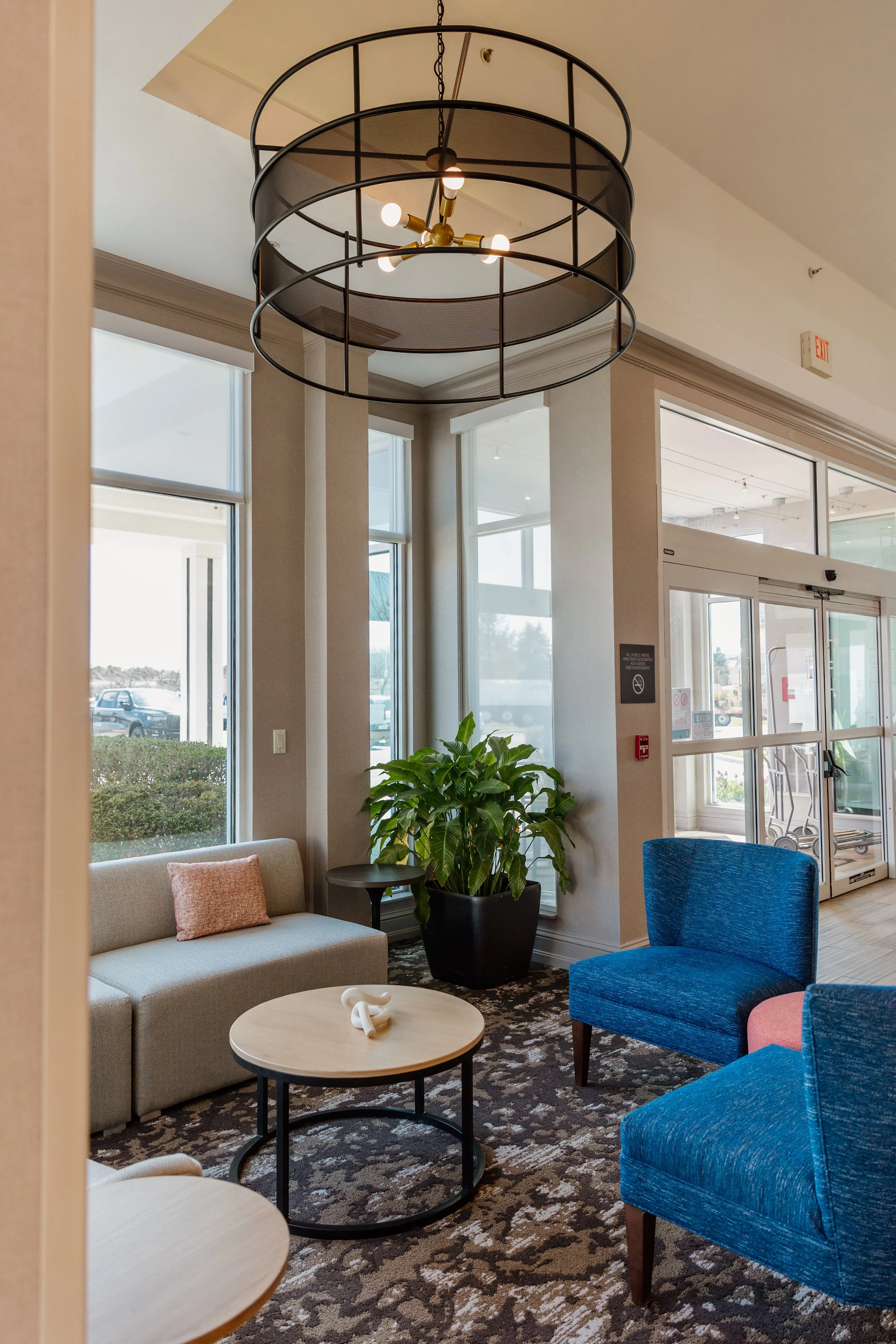 Interior of a modern lobby area featuring a black circular chandelier, a beige sofa with a pink pillow, a blue armchair, a potted plant, and round coffee tables, near large windows and a glass door.