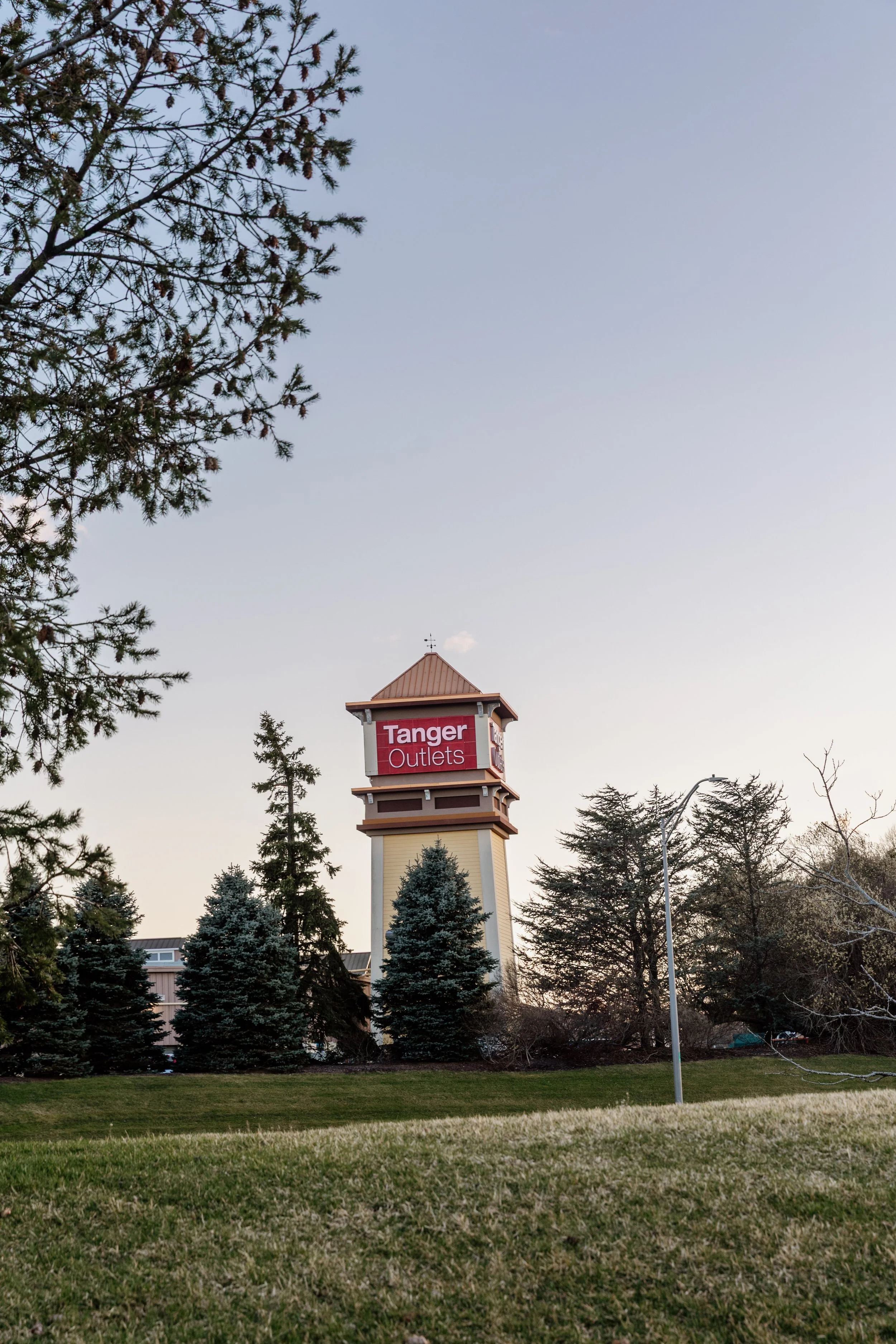 View of a water tower with a sign that reads "Tanger Outlets" during daylight, surrounded by trees and grass.
