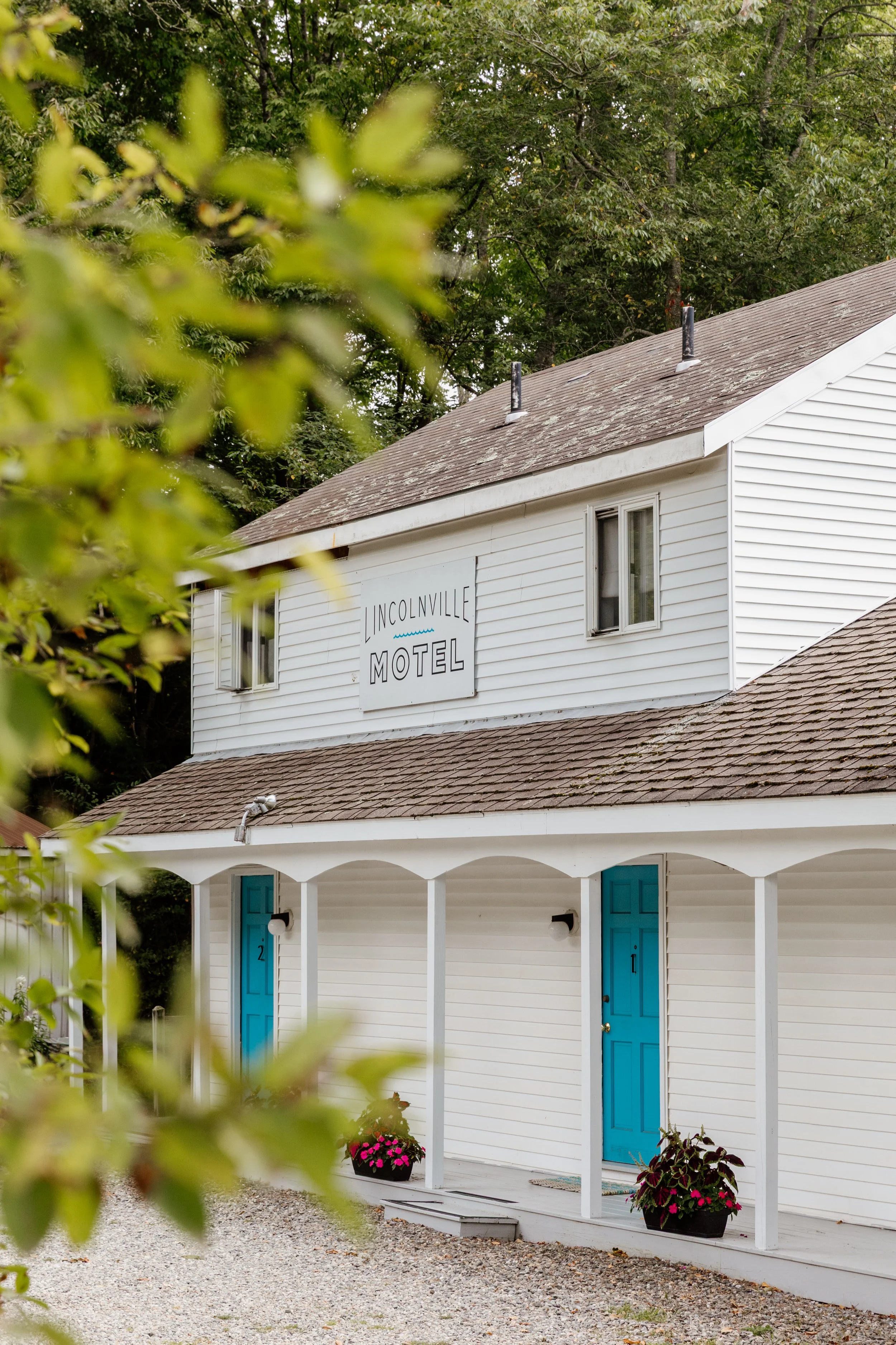 A two-story white building with a porch, blue doors, and a sign that reads "Inconville Motel". Green foliage partially obscures the view.