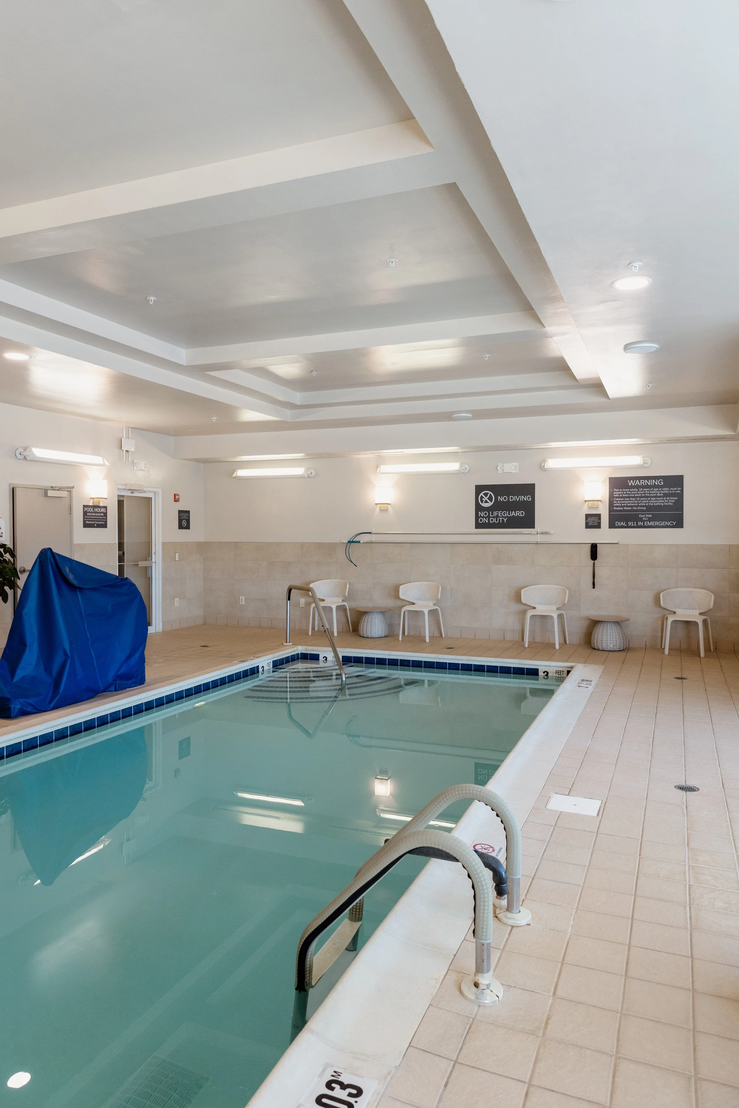 Indoor swimming pool area with a few chairs, safety signs on the wall, and a blue covered object next to a door.