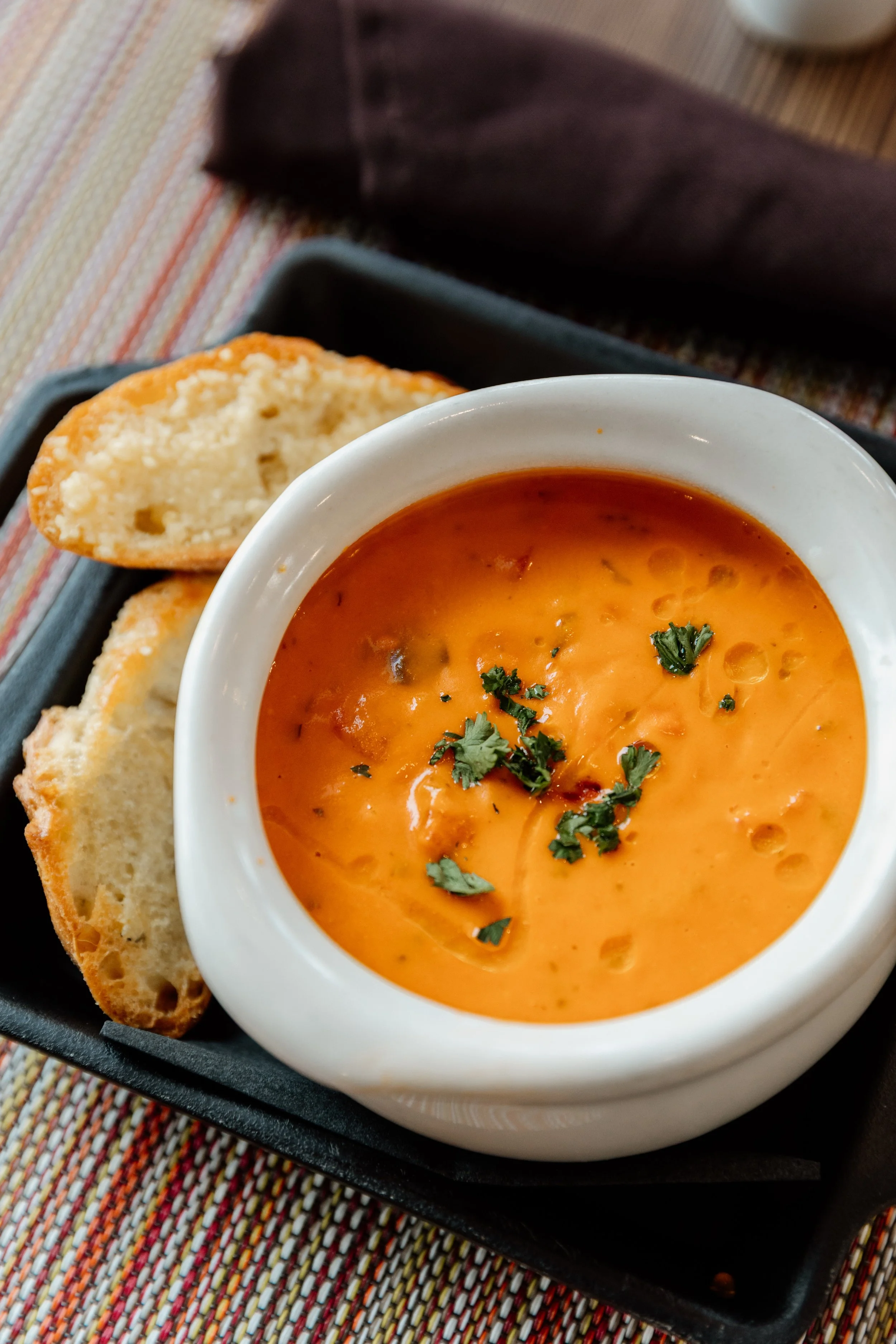 A bowl of creamy tomato soup garnished with cilantro, served with slices of toasted bread, placed on a black tray with a colorful striped tablecloth.