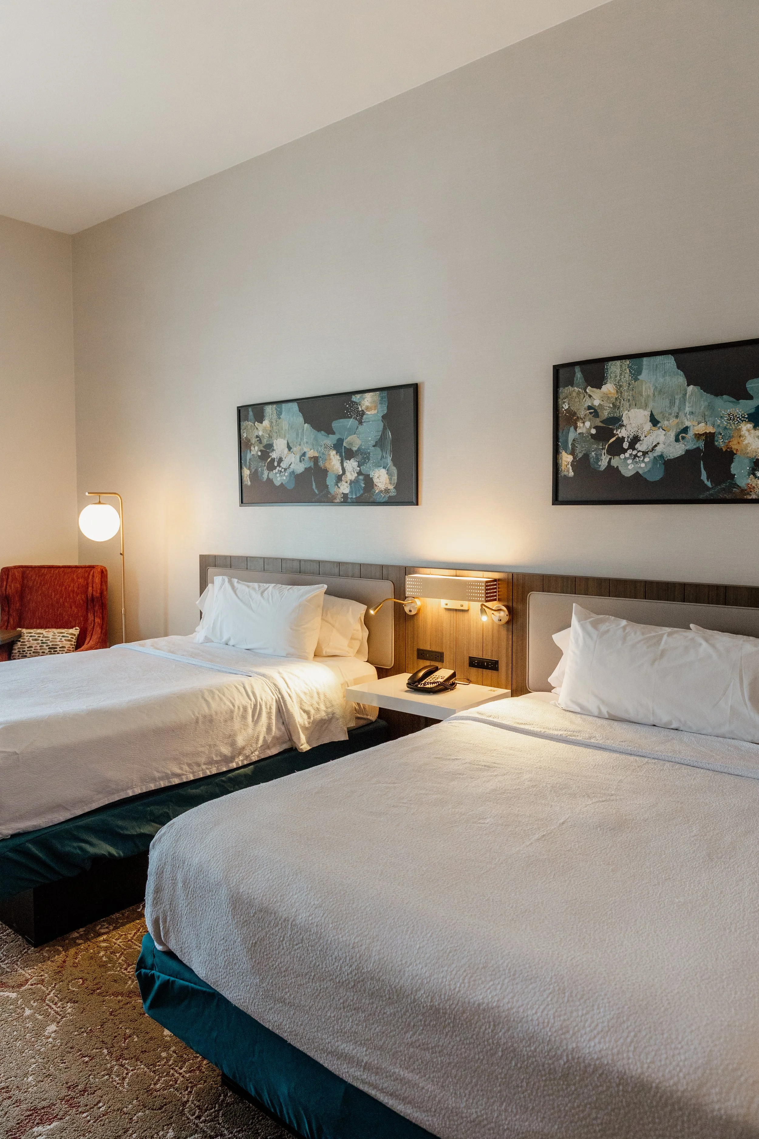 A hotel room with two beds, artwork above, a bedside table with a phone, and a lamp, with a red armchair in the corner.