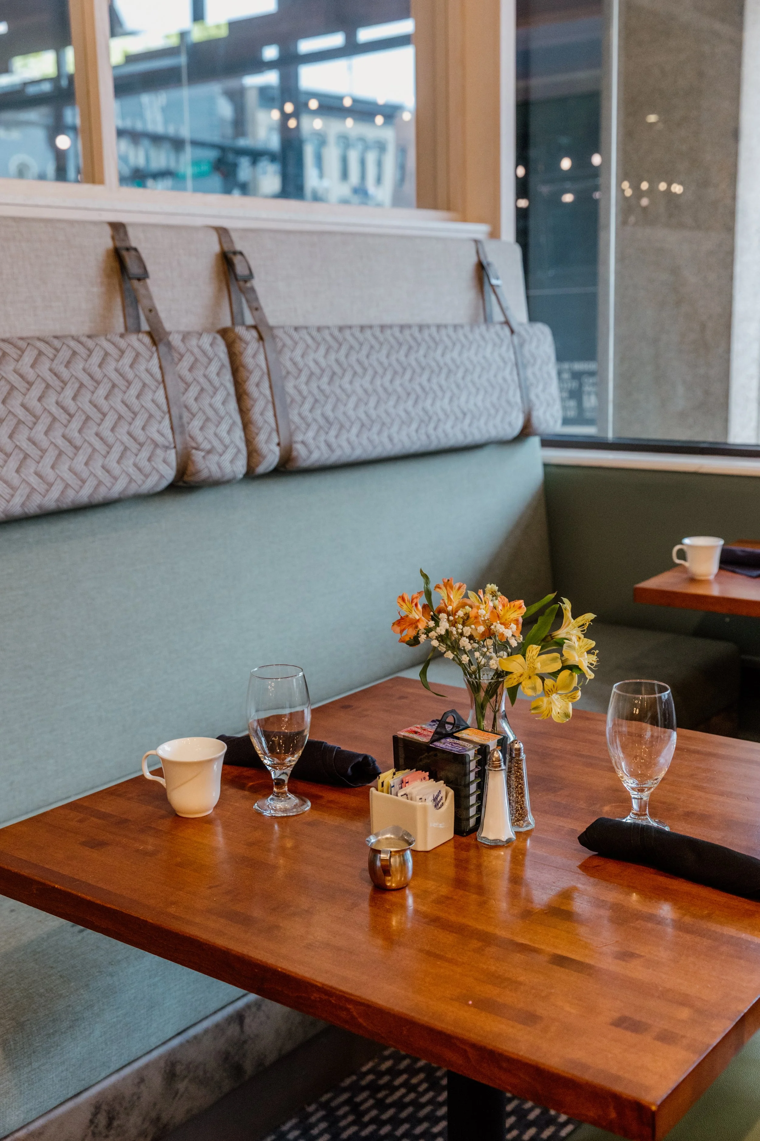 A restaurant booth with a wooden table, a green upholstered bench, and a high back with cushion straps. The table has a small vase of yellow and orange flowers, two empty wine glasses, a coffee cup, a napkin, and condiment containers.