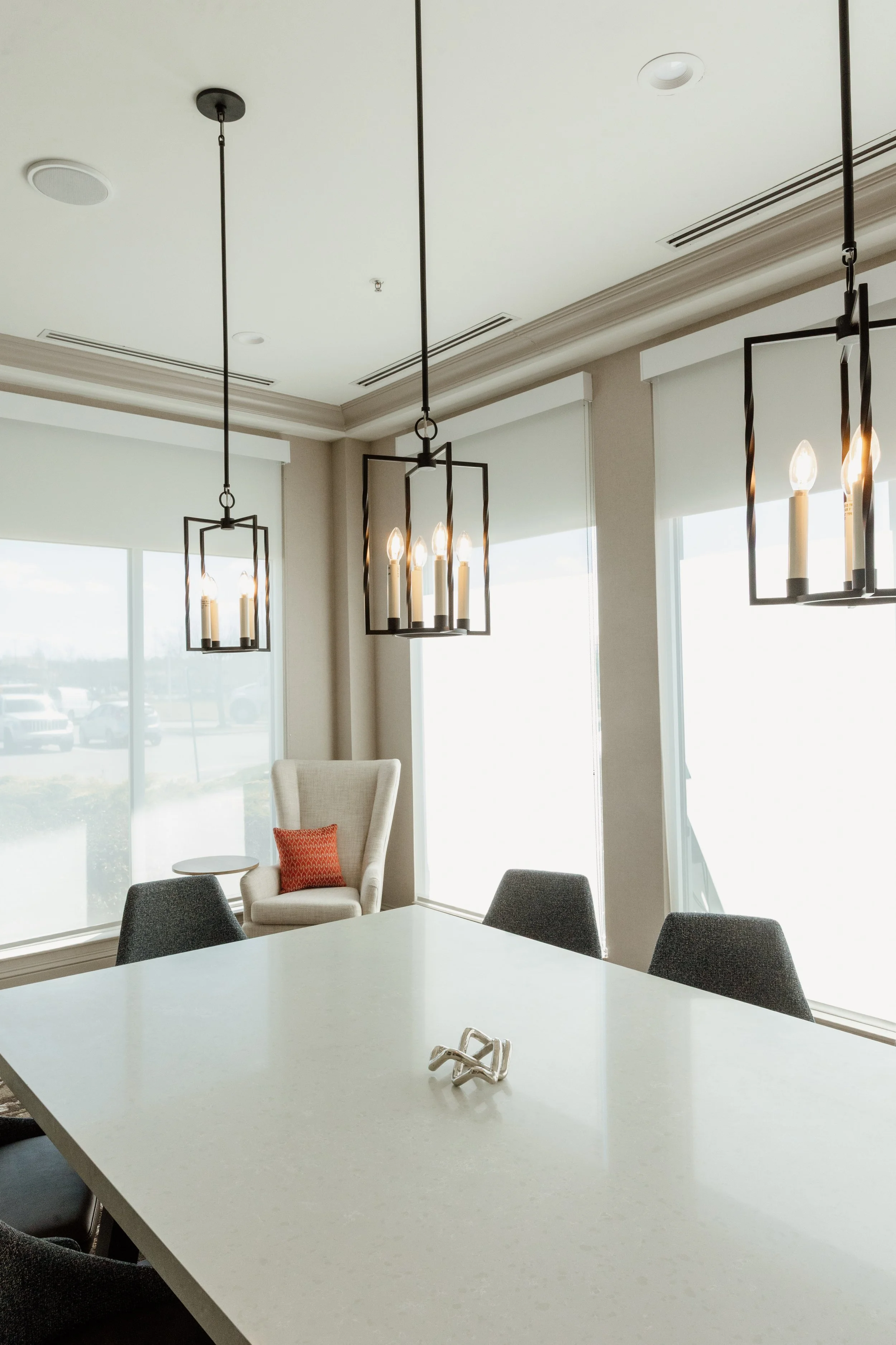 Modern conference room with a large white table, gray chairs, a high-back armchair with a red pillow, and three pendant lights with black frames and candle-like bulbs.
