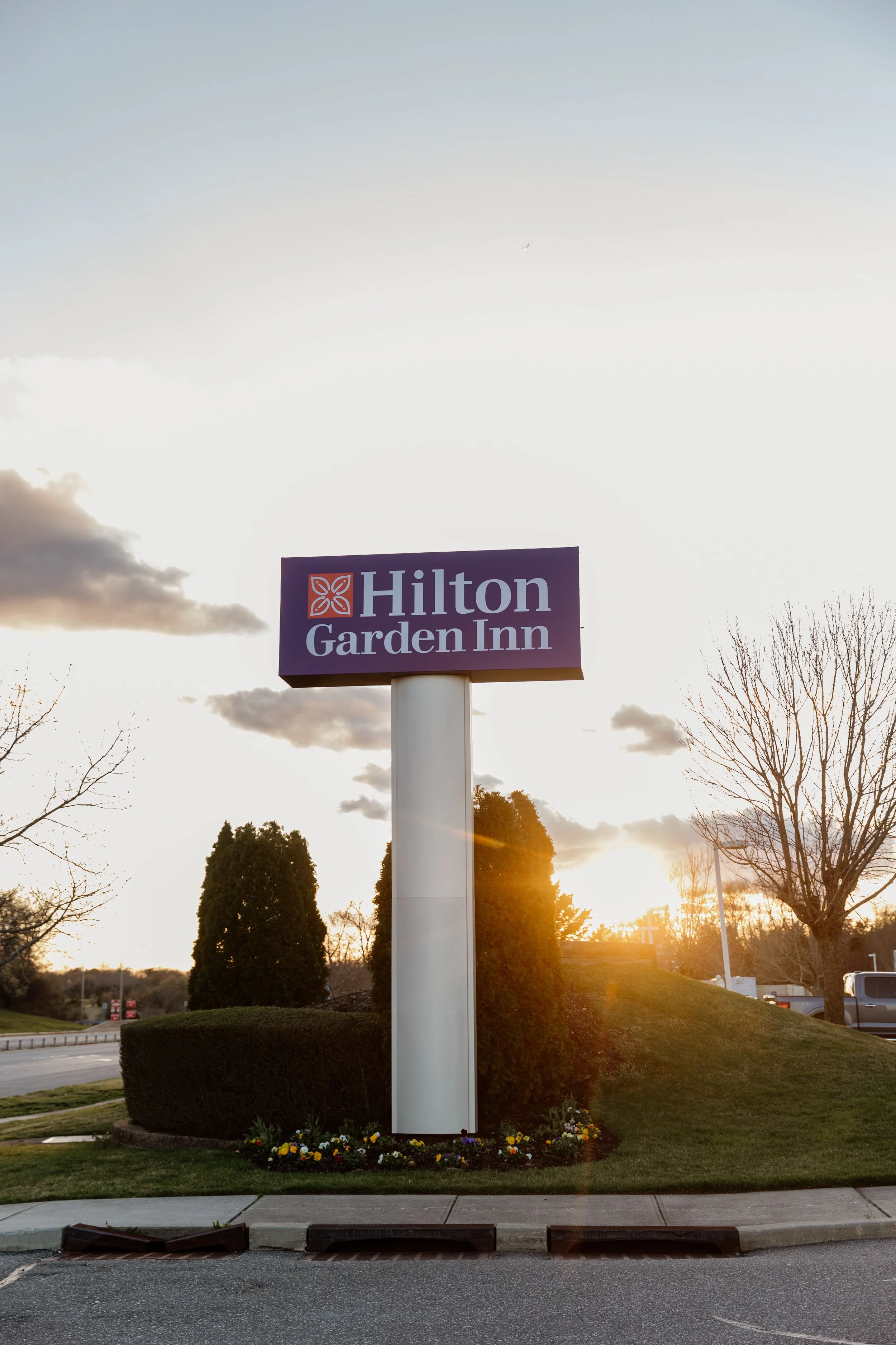 A sign for the Hilton Garden Inn located on a grassy area with trees and a parking lot in the background during sunset.