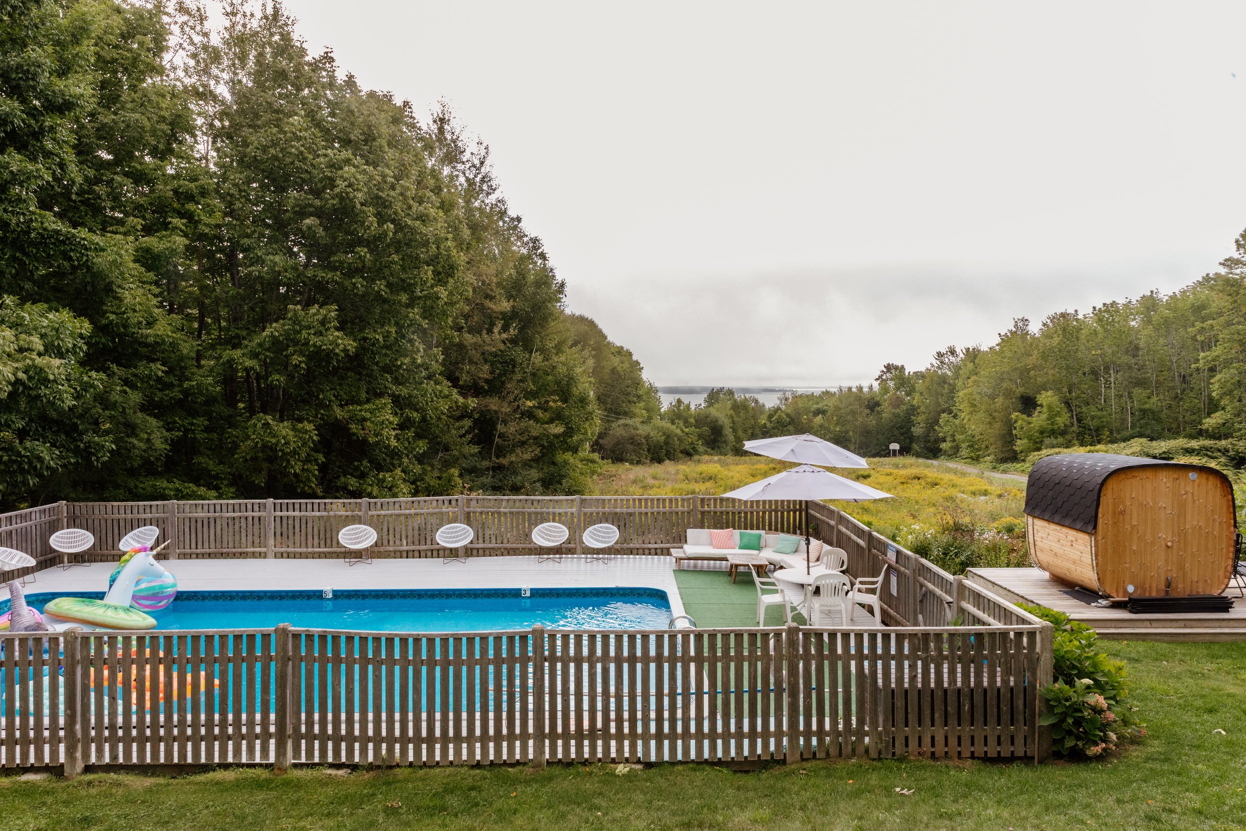 A backyard with a fenced-in pool area, pool floats, outdoor seating, umbrellas, and a small wooden sauna on a deck, surrounded by lush green trees and a cloudy sky.