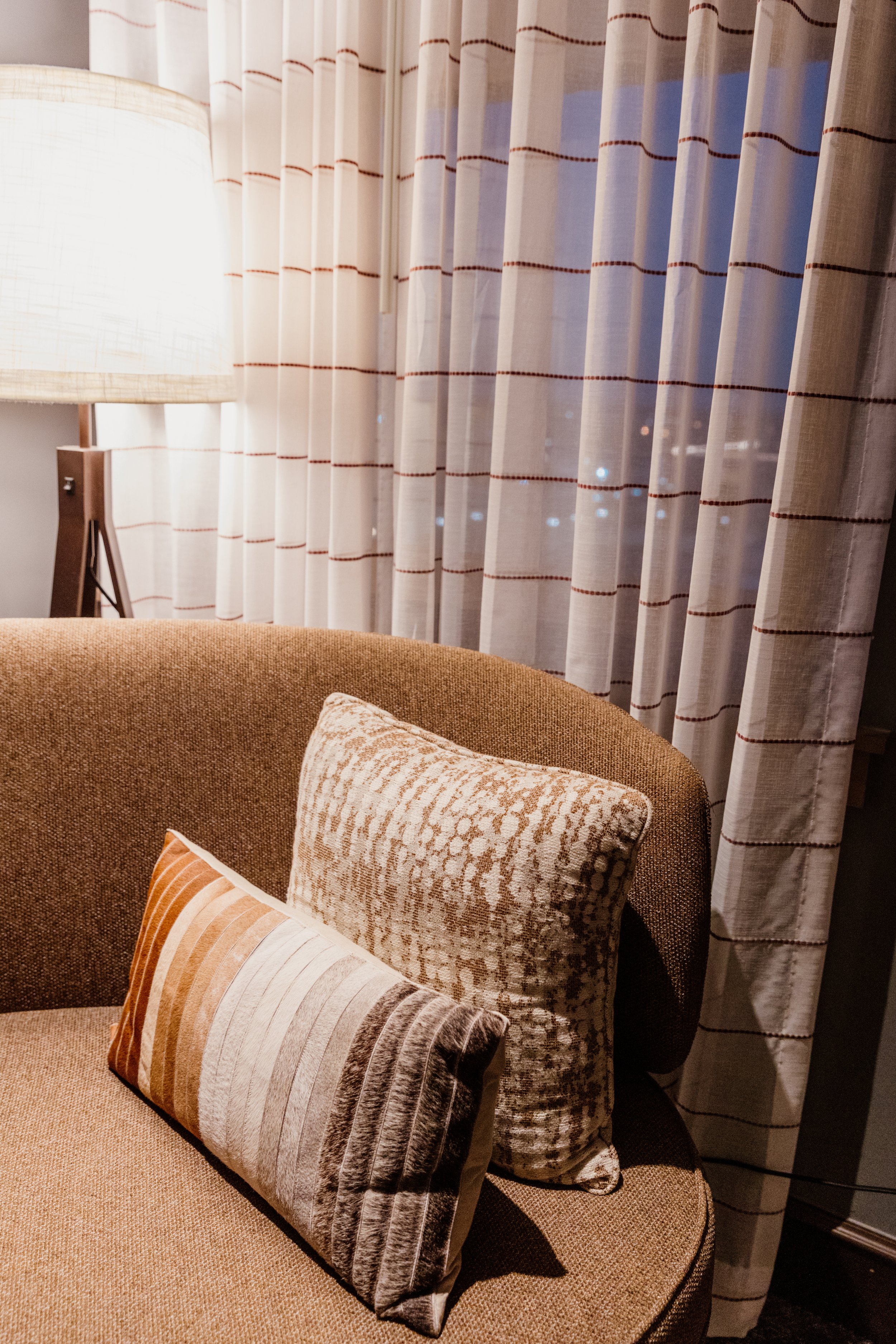 A cozy corner with a brown upholstered armchair, decorative pillows, sheer curtains, and a standing floor lamp nearby.