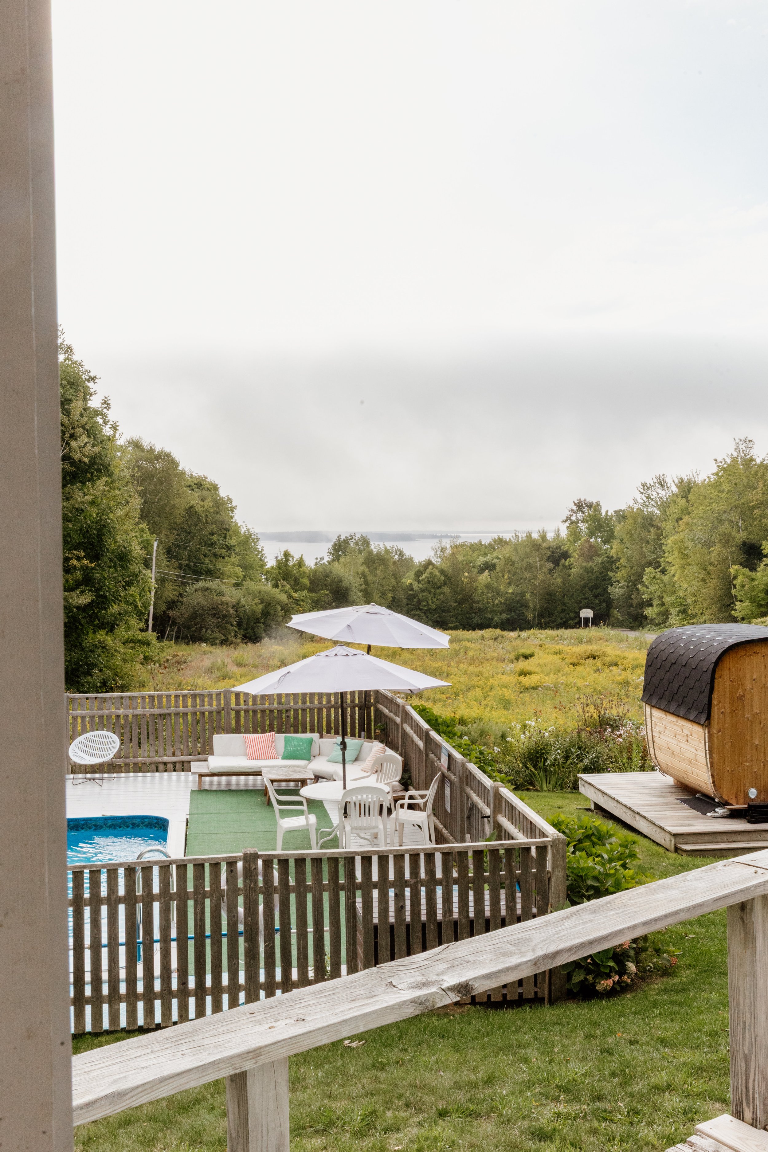 View of a backyard deck with white patio furniture, umbrellas, and a small pool, overlooking a grassy field, trees, and a body of water in the distance on a cloudy day.
