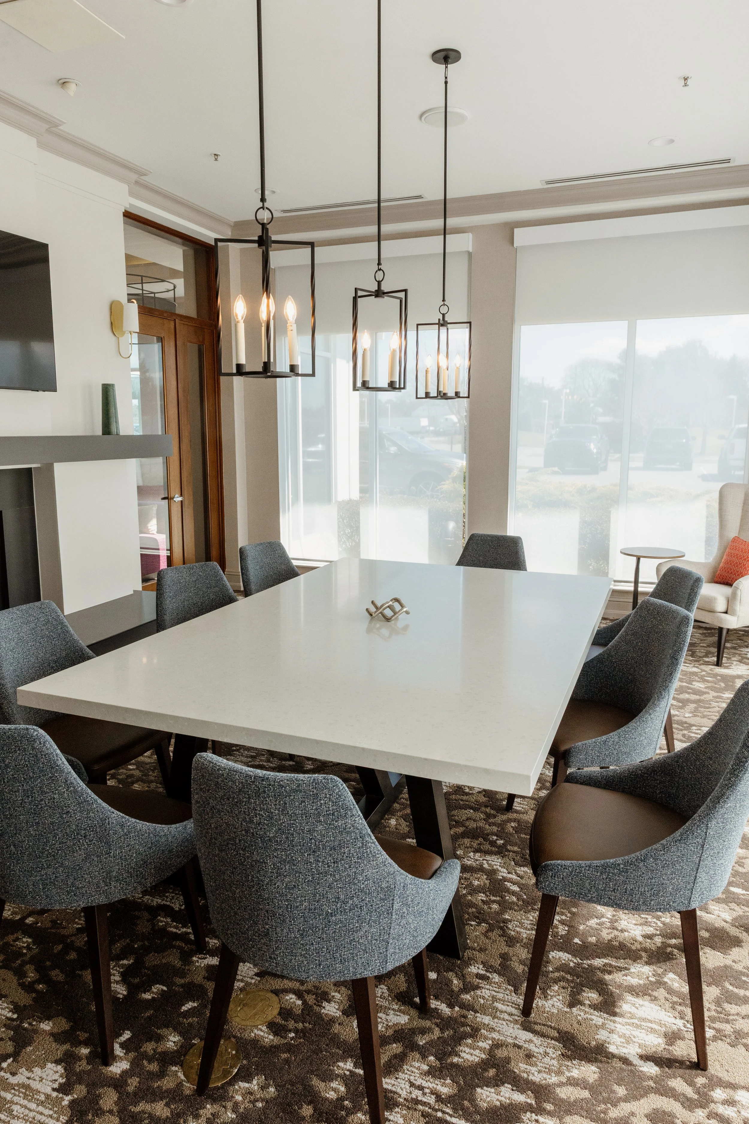 A modern conference room featuring a large white table surrounded by eight dark grey upholstered chairs. Elegant chandelier lighting hangs above the table, and large windows let in natural light. A patterned carpet covers the floor, and a beige armch