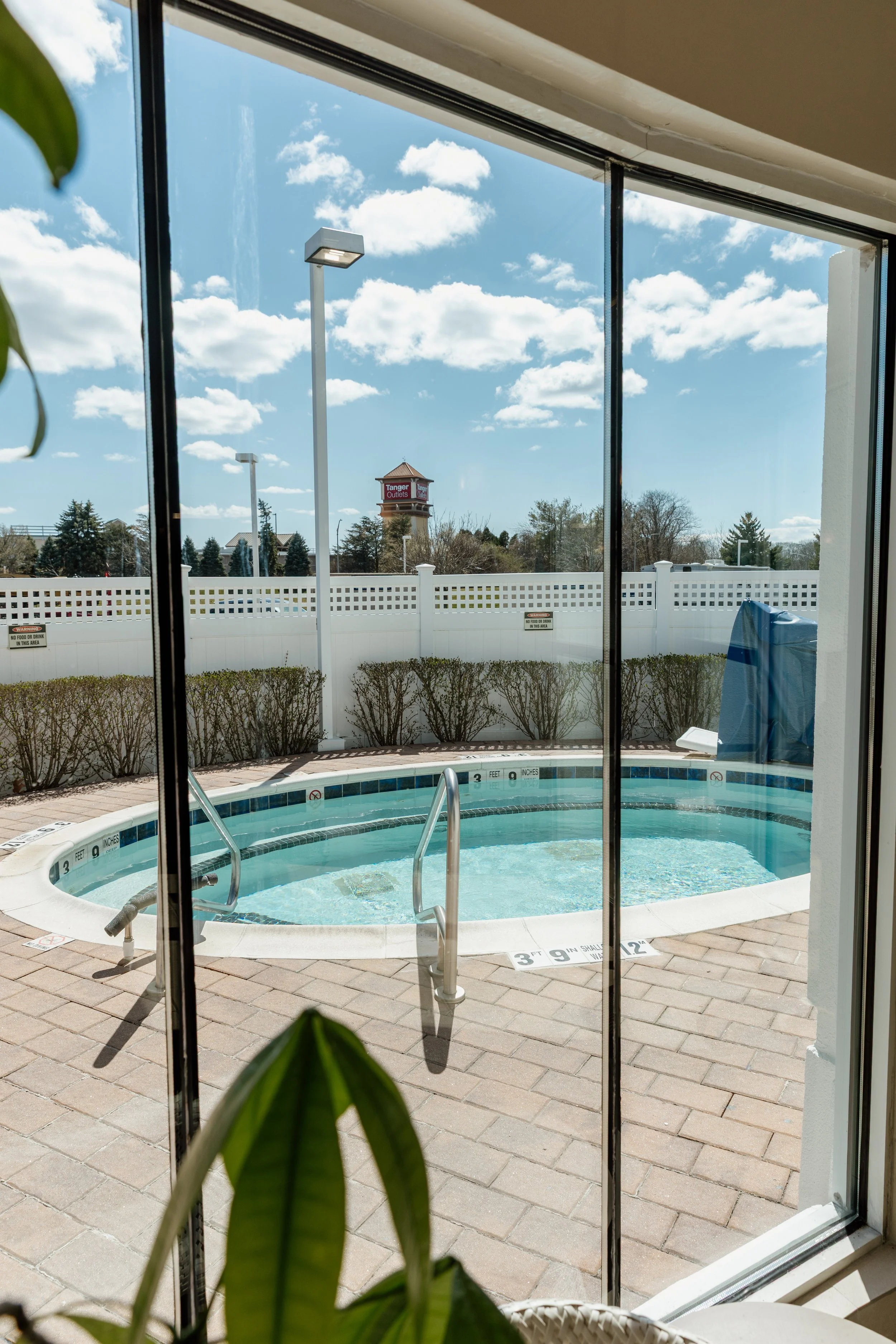 View of a small outdoor hot tub or spa seen through glass sliding doors, with clear blue sky and some clouds in the background, a white fence surrounding the area, and a few bushes along the fence line.