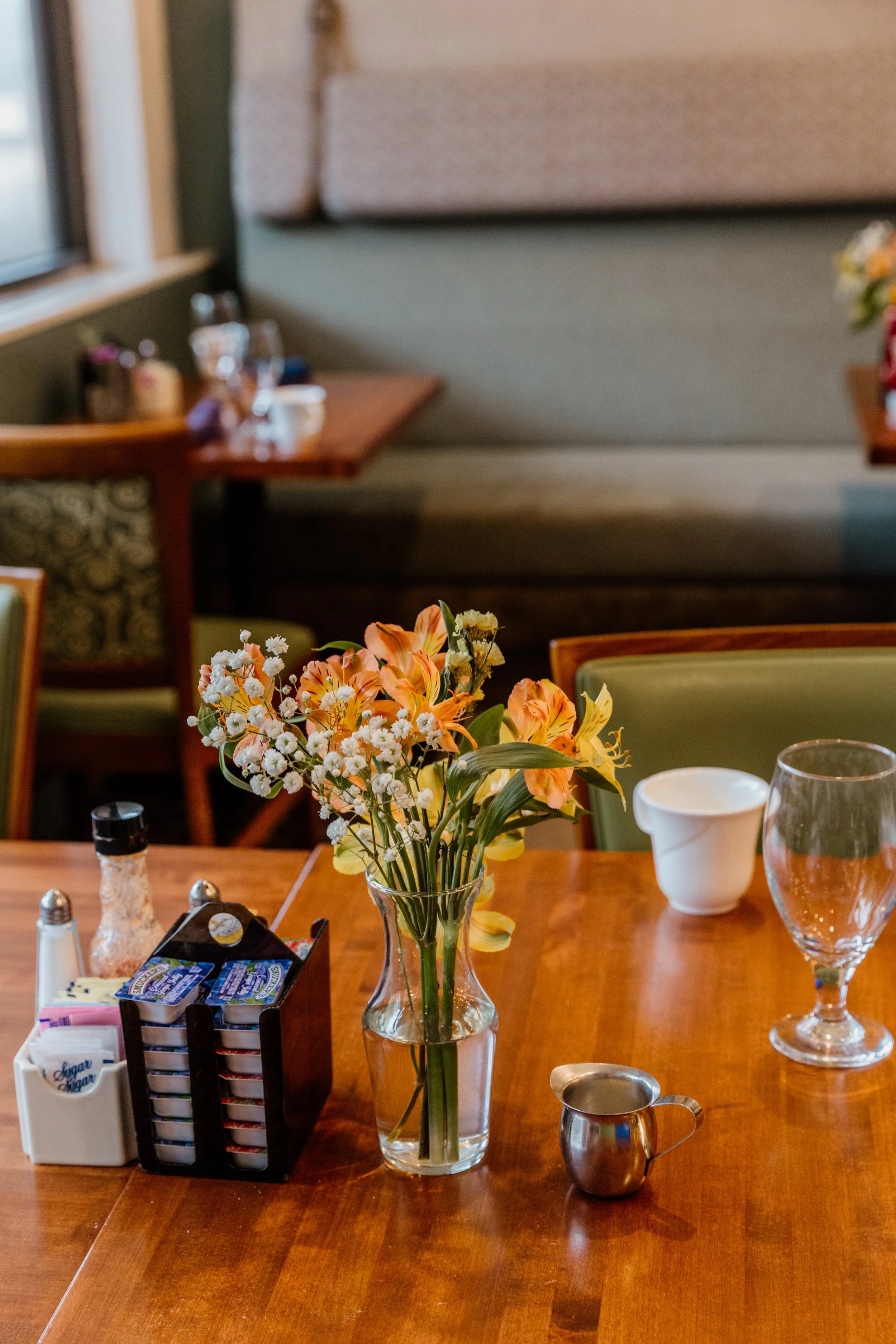 A restaurant table set with a glass vase of orange and yellow flowers, a glass, a small silver creamer, and condiments, with other tables visible in the background.