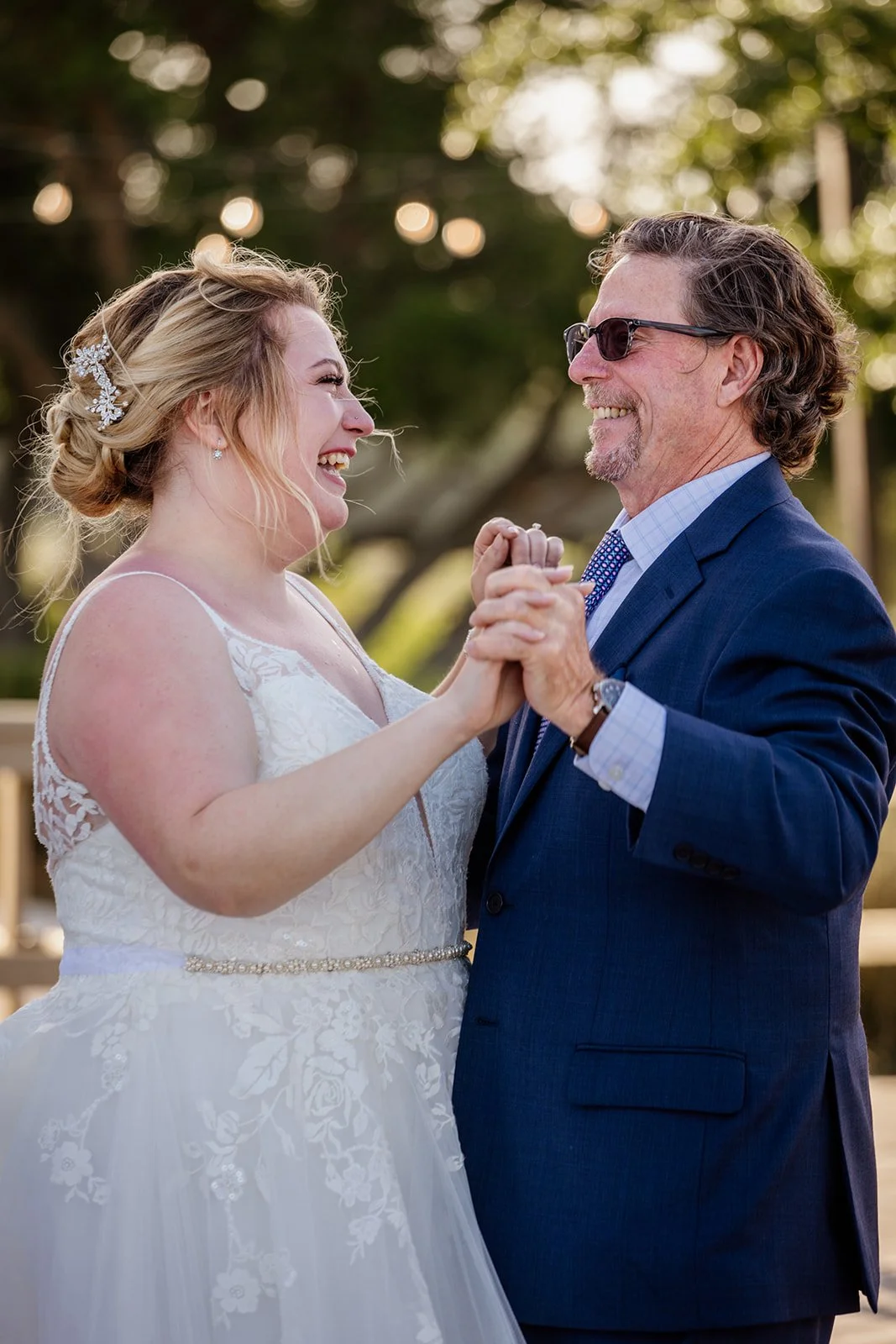 A bride and an older man, possibly her father, are dancing and smiling at an outdoor wedding. The bride wears a white lace wedding gown with a beaded belt and a floral hairpiece. The man is dressed in a blue suit, light blue shirt, tie, and sunglasses.