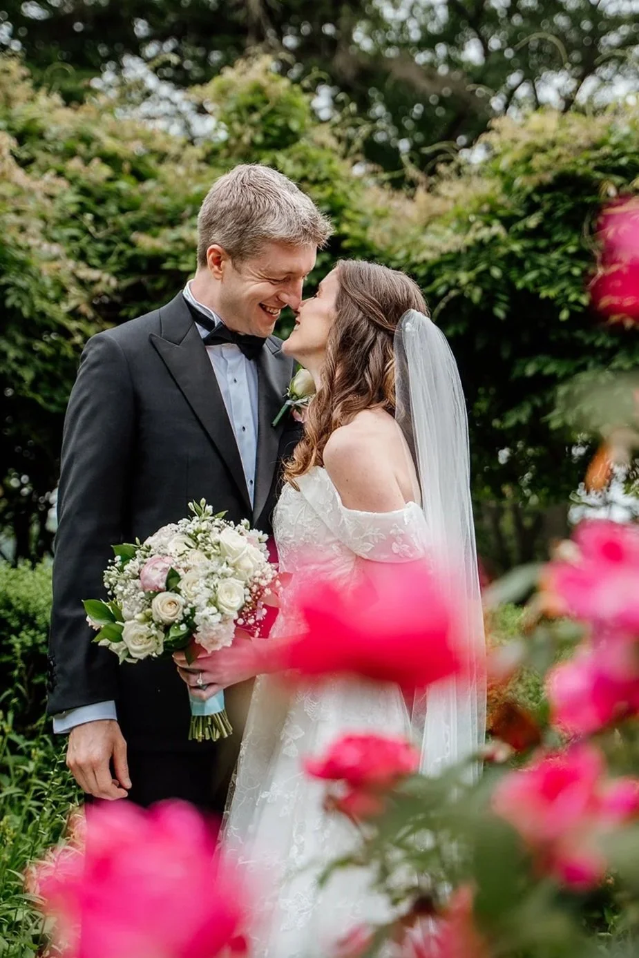 A bride and groom leaning in close and sharing a smile in the rose garden at Saint Clements Castle and Marina in Portland, Connecticut, surrounded by pink blooms, the bride holding a bouquet of white and pink roses.