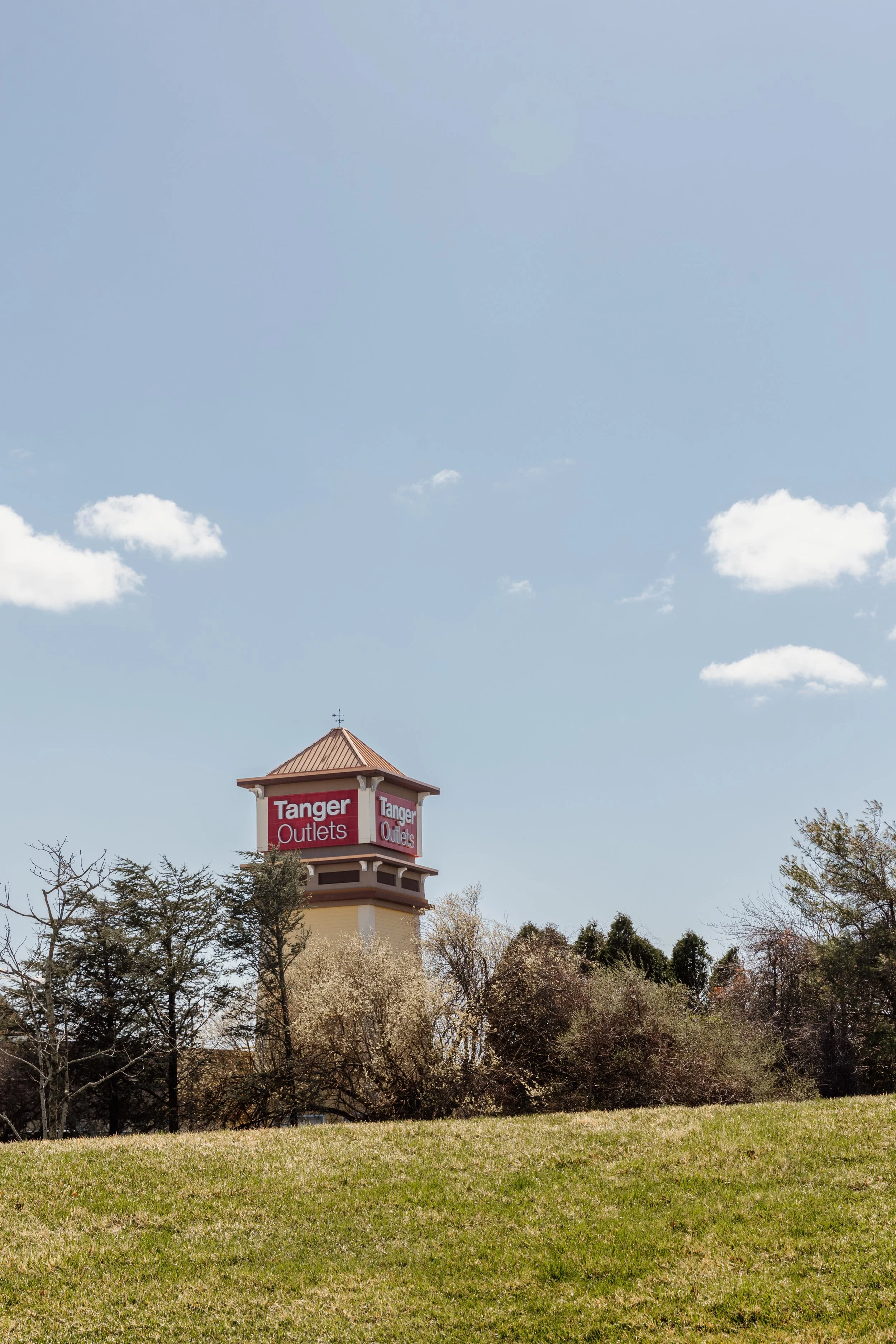 A water tower with red signage that reads "Tanger Outlets" in the distance, surrounded by trees and an open grassy area under a partly cloudy sky.