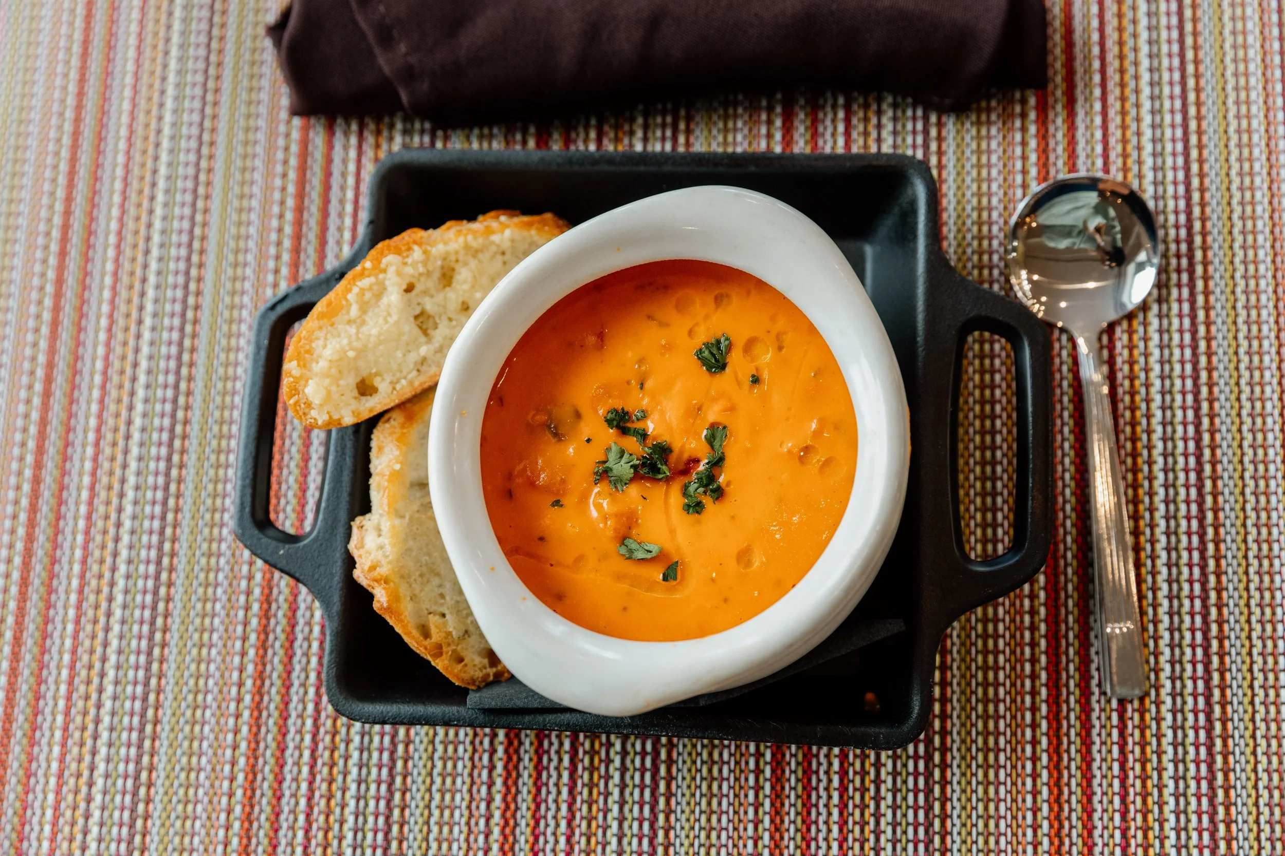 Bowl of orange soup garnished with chopped herbs, served with slices of bread on a black tray, with a spoon beside it on a colorful striped tablecloth.