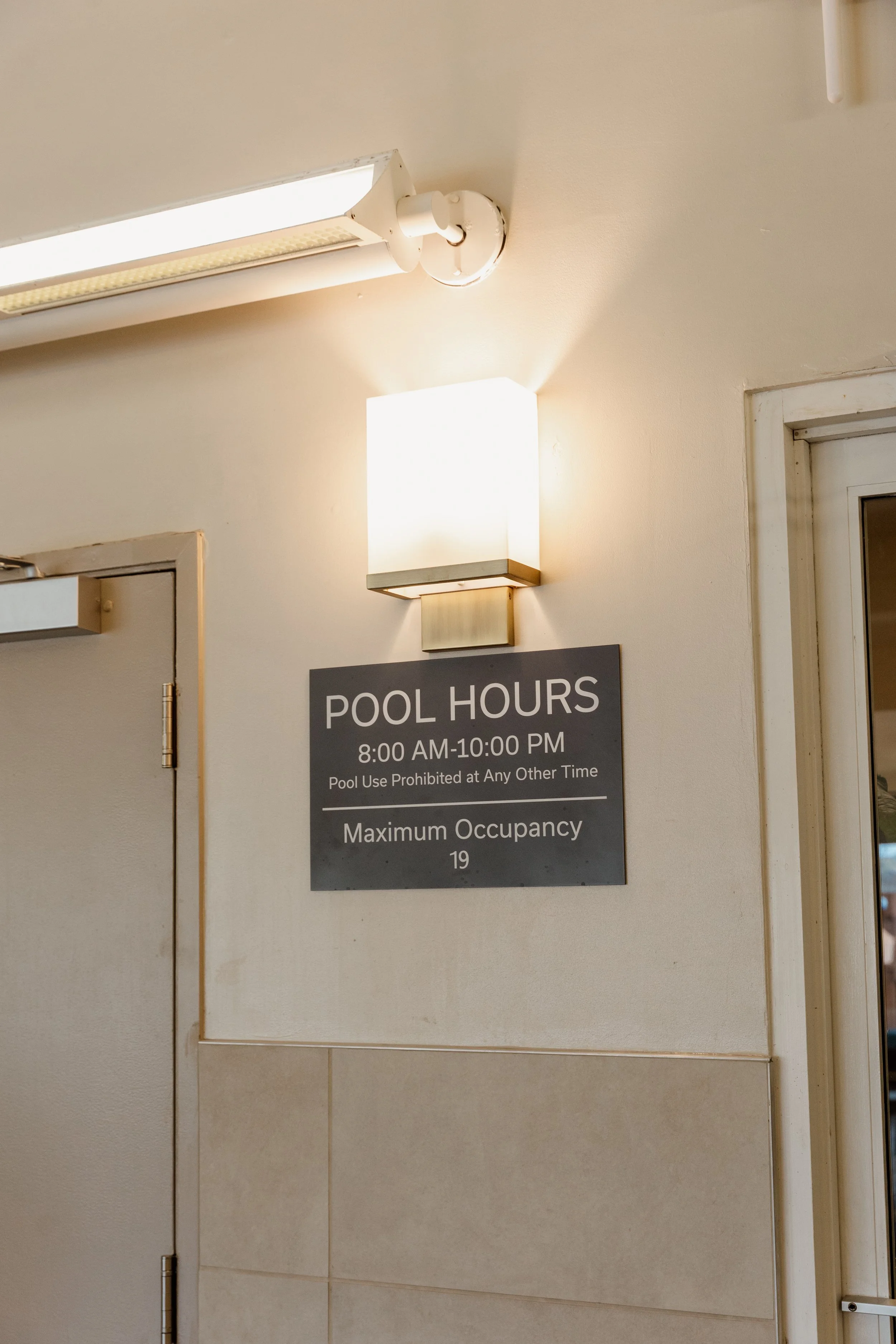 A sign displaying pool hours from 8:00 AM to 10:00 PM, with a maximum occupancy of 19, mounted on a wall next to a light fixture.