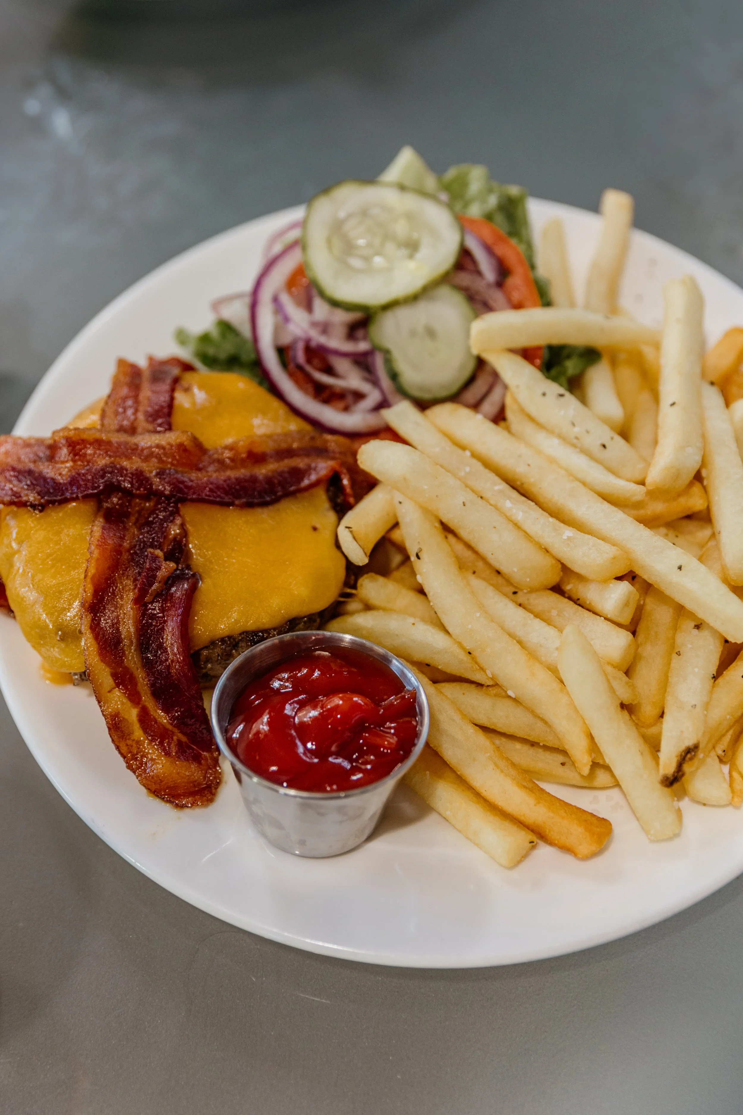 Plate of cheeseburger with bacon, French fries, a side of ketchup, and a small salad with cucumbers, tomatoes, and red onions.