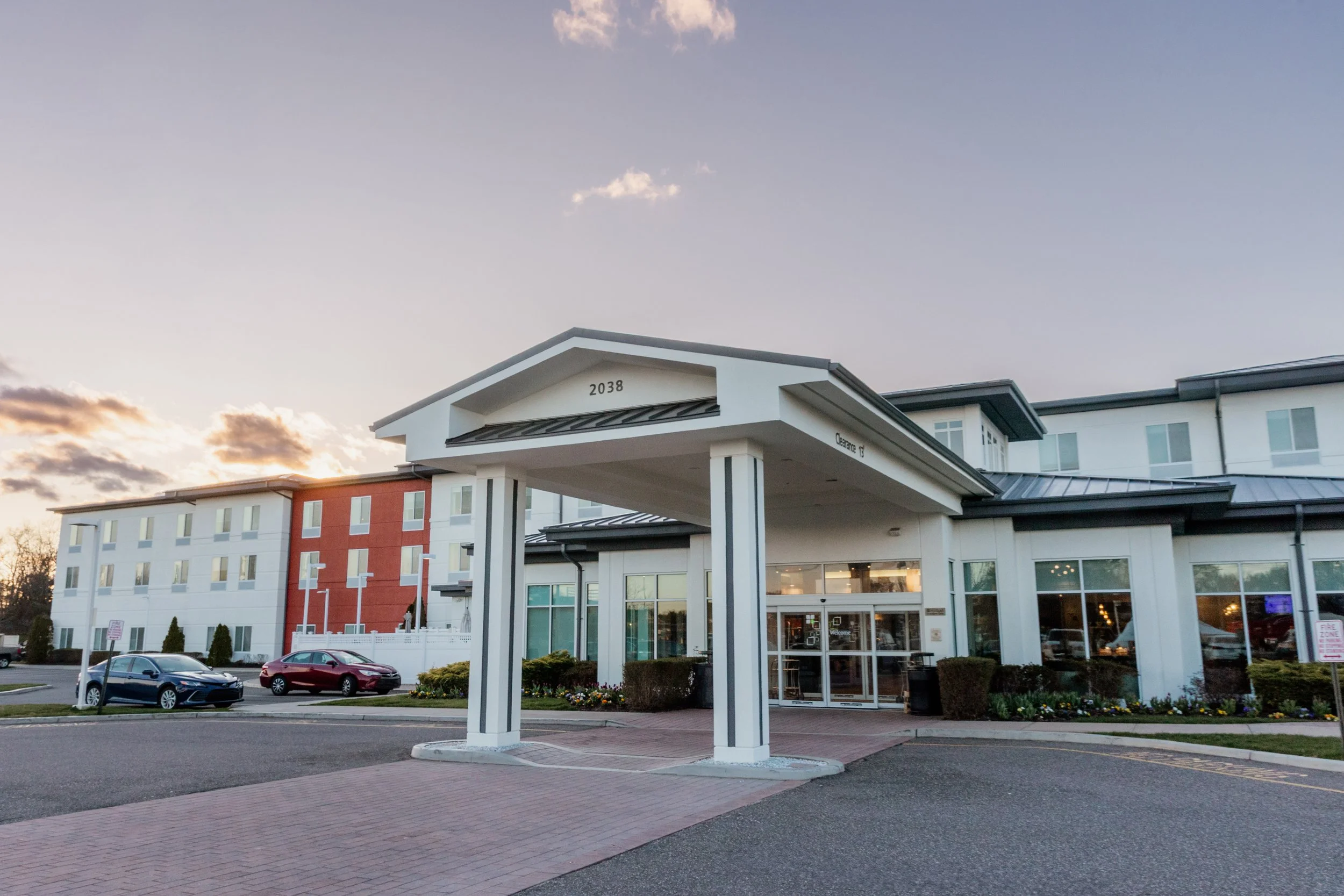 Exterior view of a modern hotel with white walls and a covered entrance, with cars parked nearby at sunset.