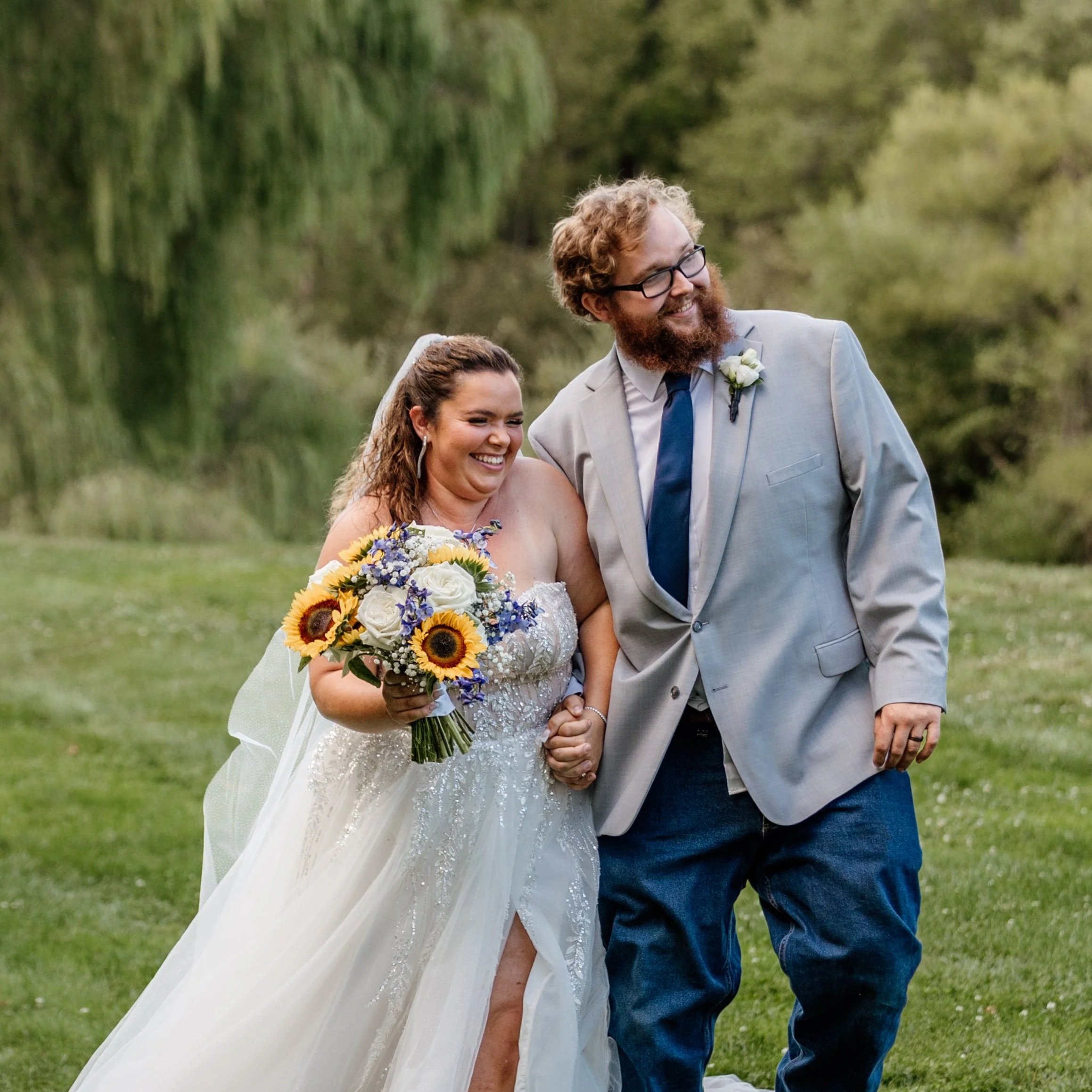 A newlywed couple walking outdoors in a park, smiling and holding hands. The bride is holding a bouquet of sunflowers, white roses, and purple flowers. The groom is wearing a light gray suit with a blue tie and has curly hair and a beard.