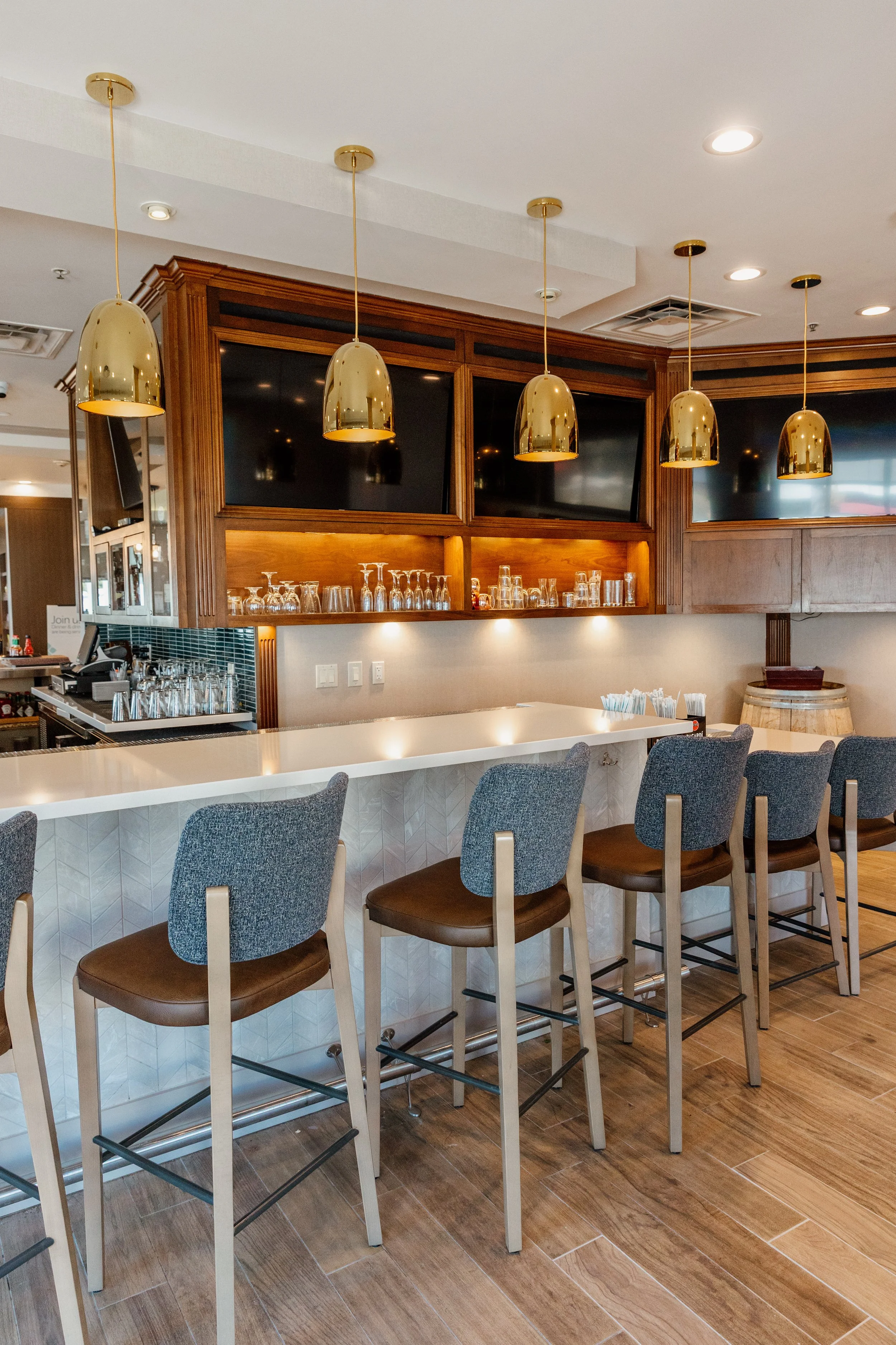 Interior of a modern bar with a white countertop, gray barstools, wooden cabinets, and gold pendant lights.