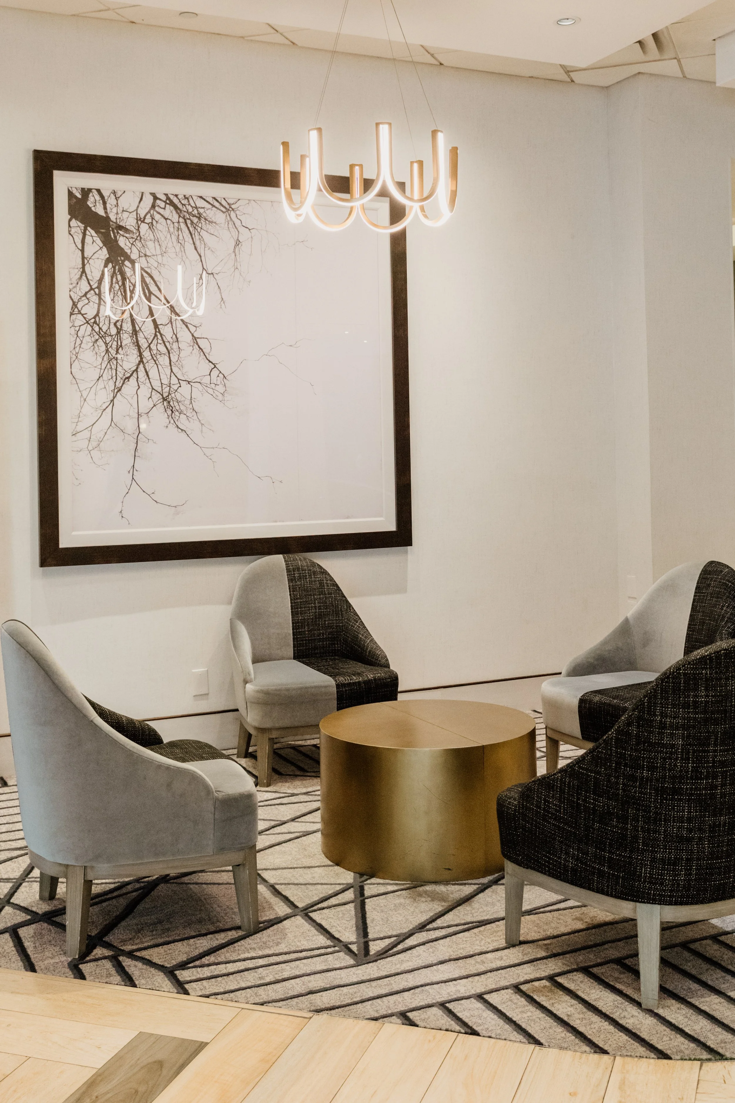 Modern hotel lobby seating area with four armchairs, a round gold coffee table, a large framed black and white picture of tree branches, and a contemporary chandelier overhead.