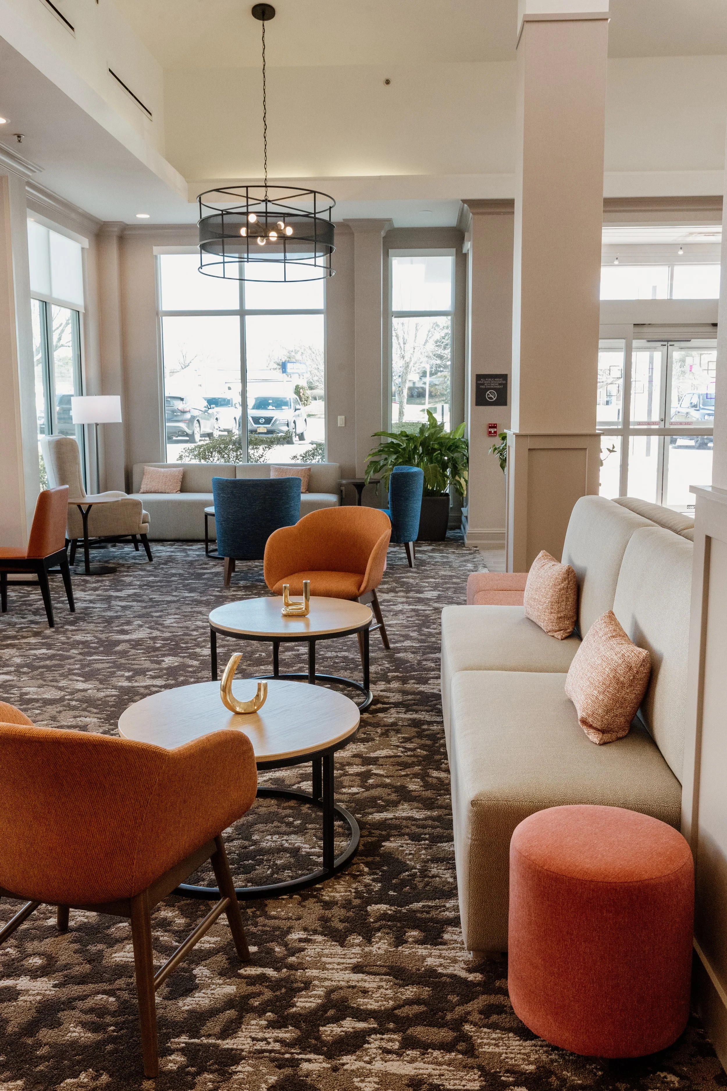 A spacious hotel lobby with large windows letting in natural light, dotted with colorful chairs and sofas, round tables, indoor plants, and a patterned carpet.