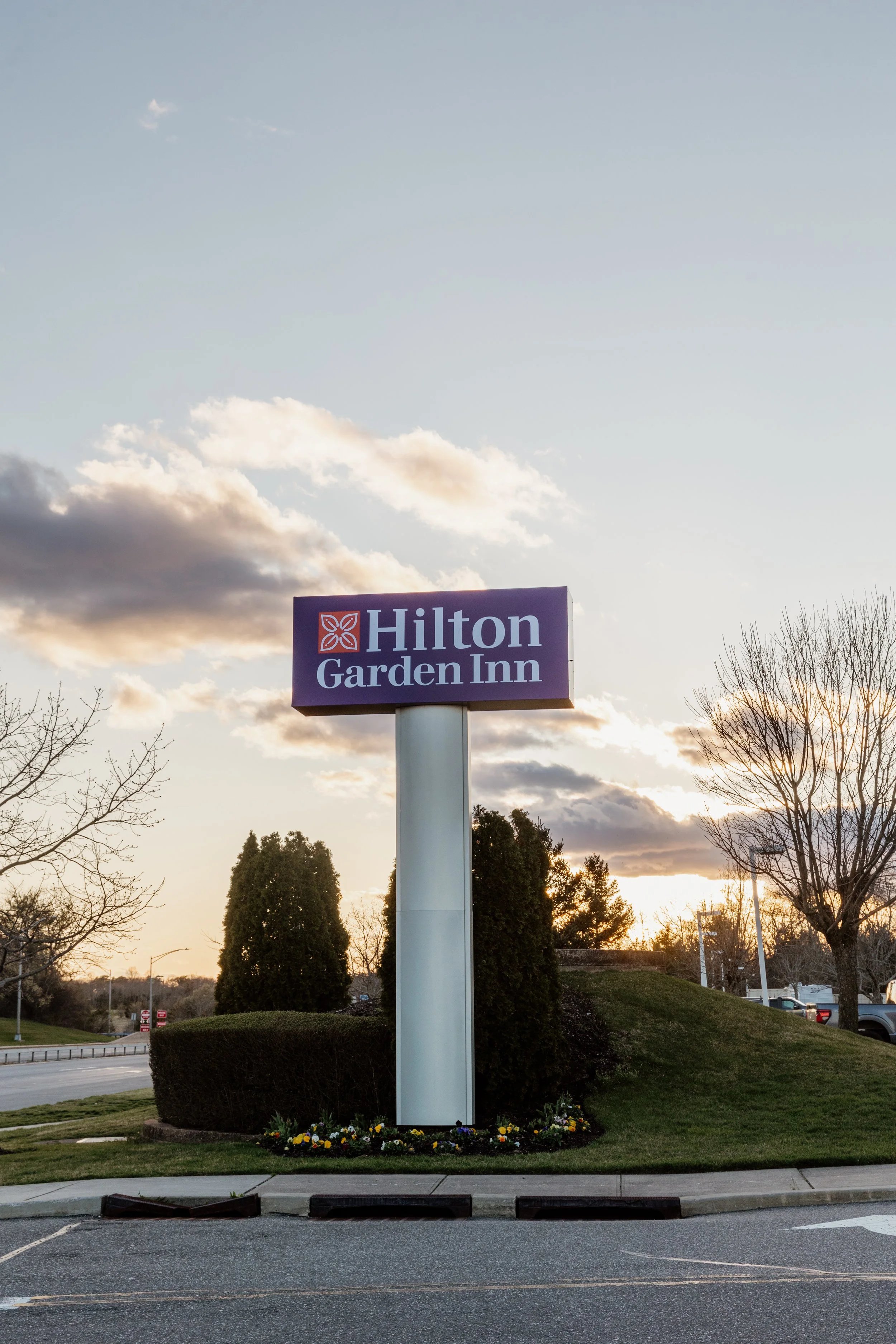 Signboard for Hilton Garden Inn hotel in a parking lot with trees and a sunset sky in the background.