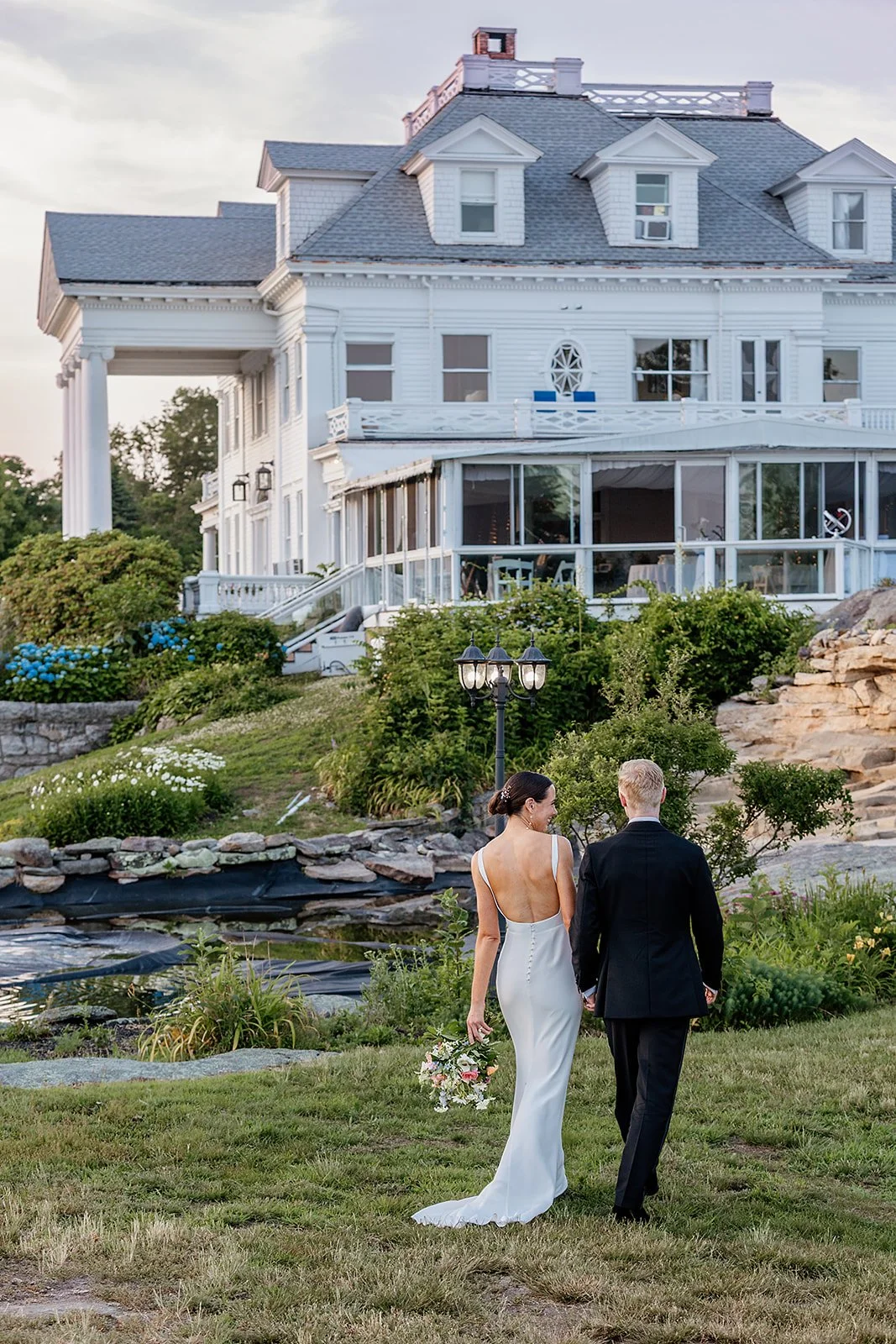 A bride and groom walking hand in hand at sunset with the stunning white facade of Haley Mansion reflected in the pond behind them in Mystic, Connecticut.