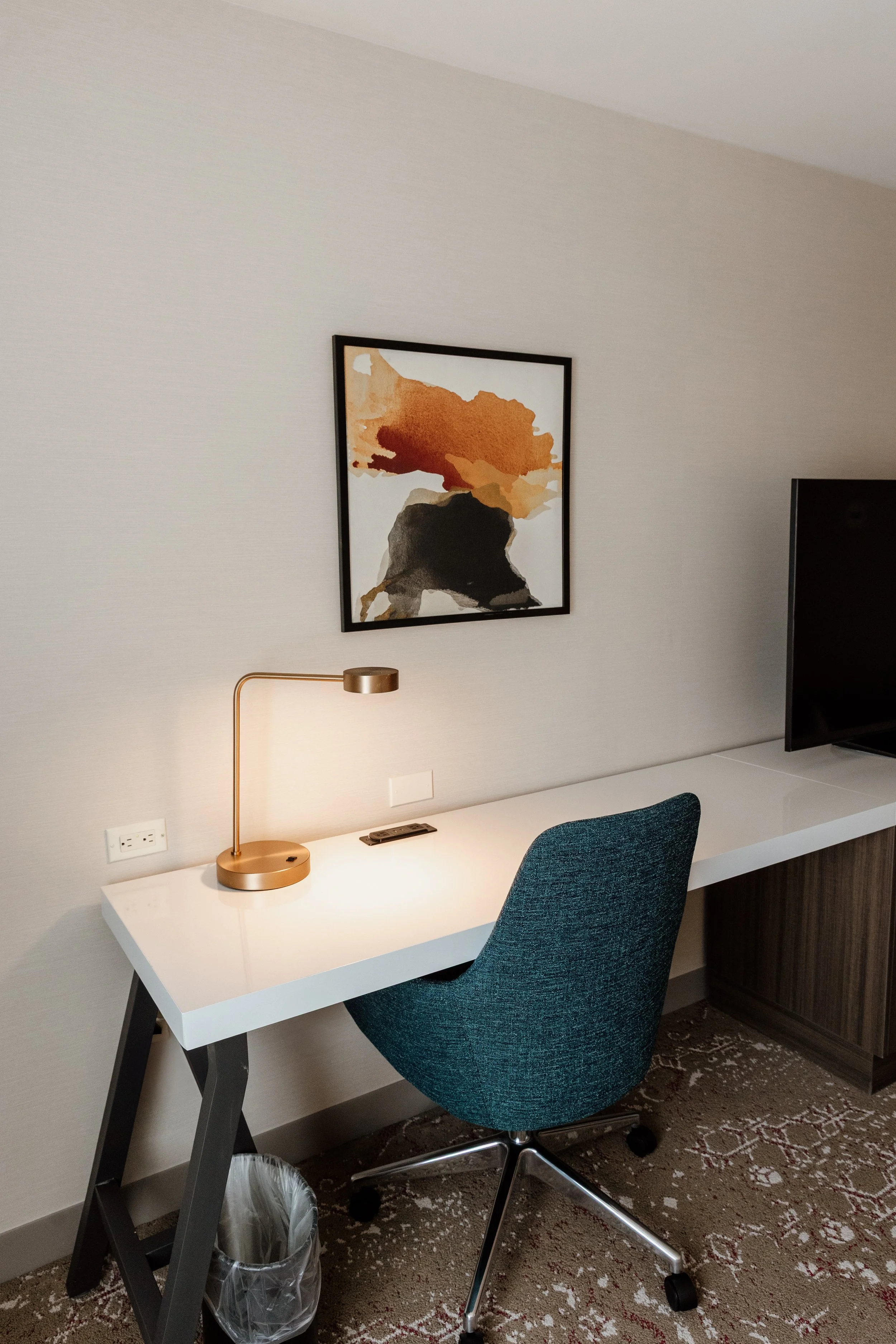 A modern hotel room workspace with a white desk, a teal office chair, a gold desk lamp, a wall outlet, a remote, artwork of colorful abstract figure, and a flat-screen television.
