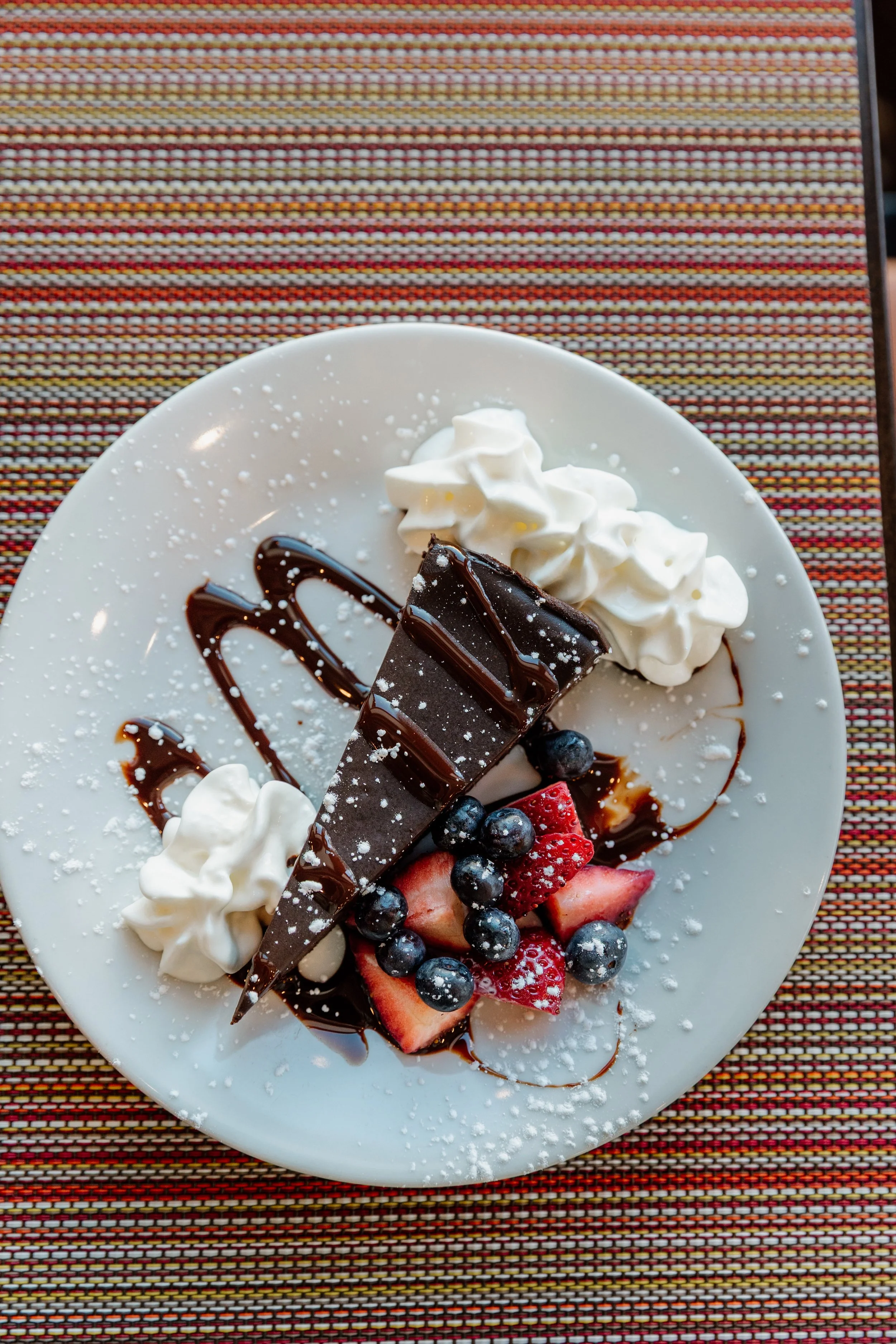 Chocolate cheesecake topped with chocolate drizzle, whipped cream, strawberries, blueberries, and powdered sugar on a white plate
