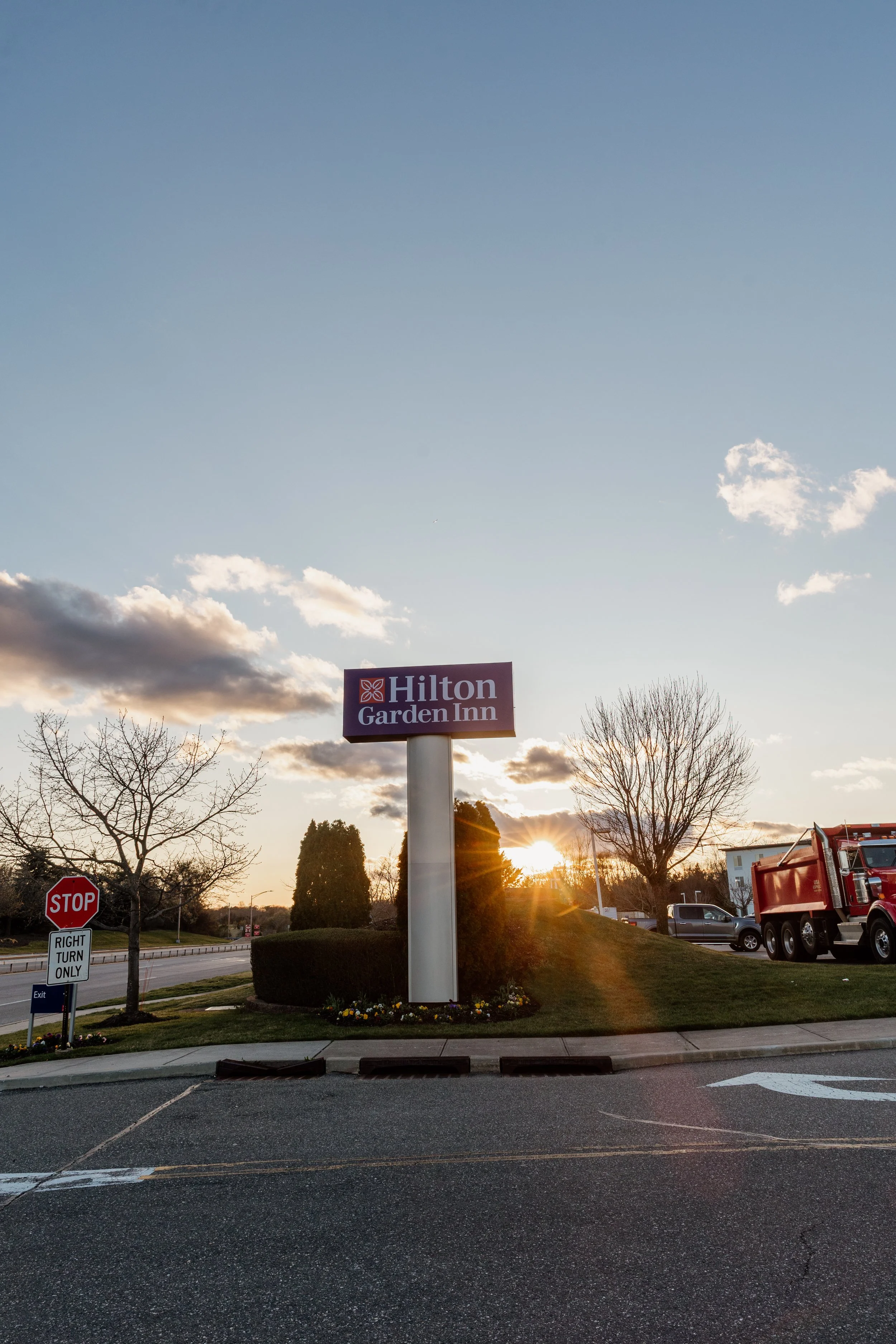 Sunset over a Hilton Garden Inn sign in a parking lot, with trees, a stop sign, and a red truck nearby.