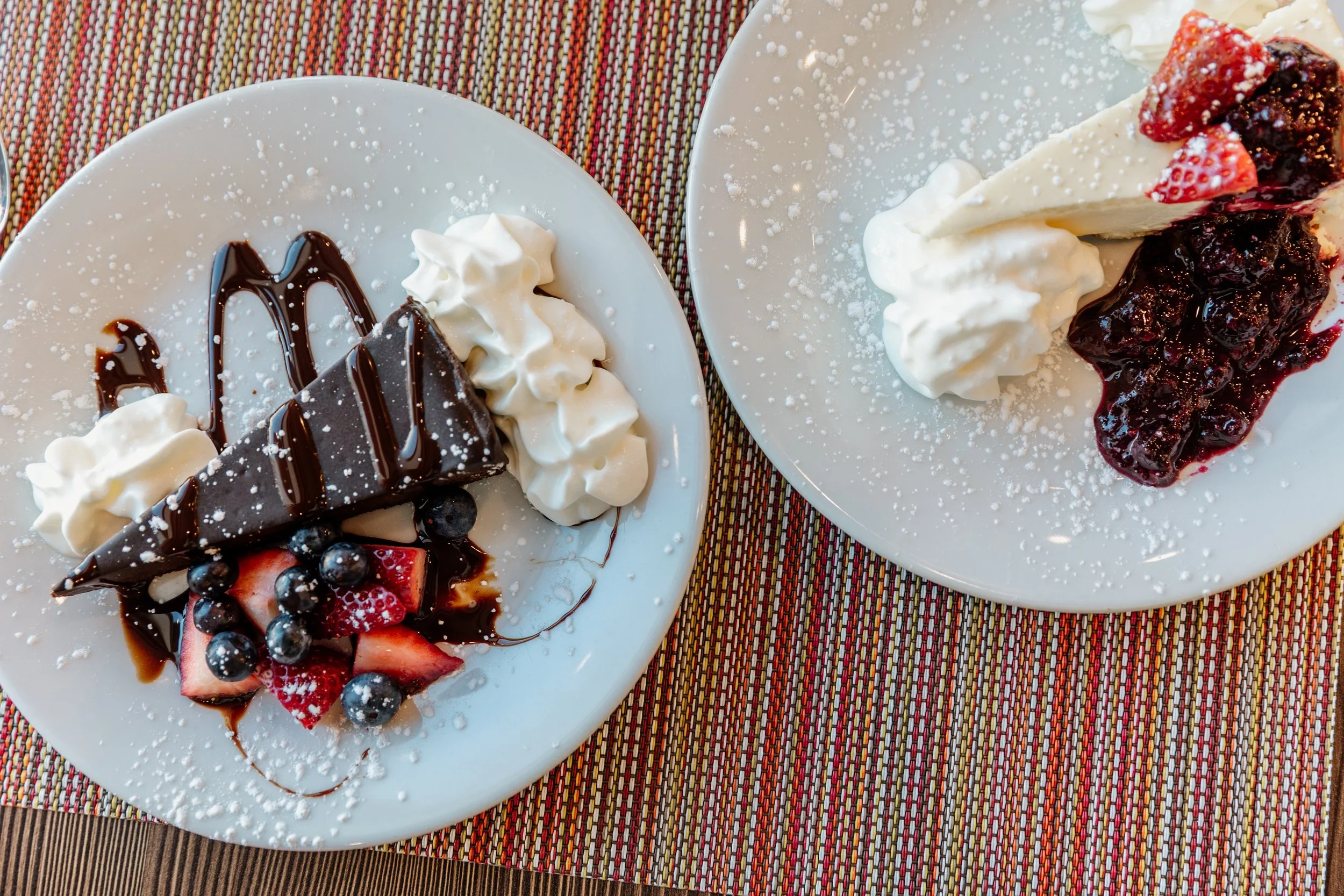 Two plates of strawberry and blueberry cheesecake with whipped cream, drizzled with chocolate sauce, on a colorful woven tablecloth.