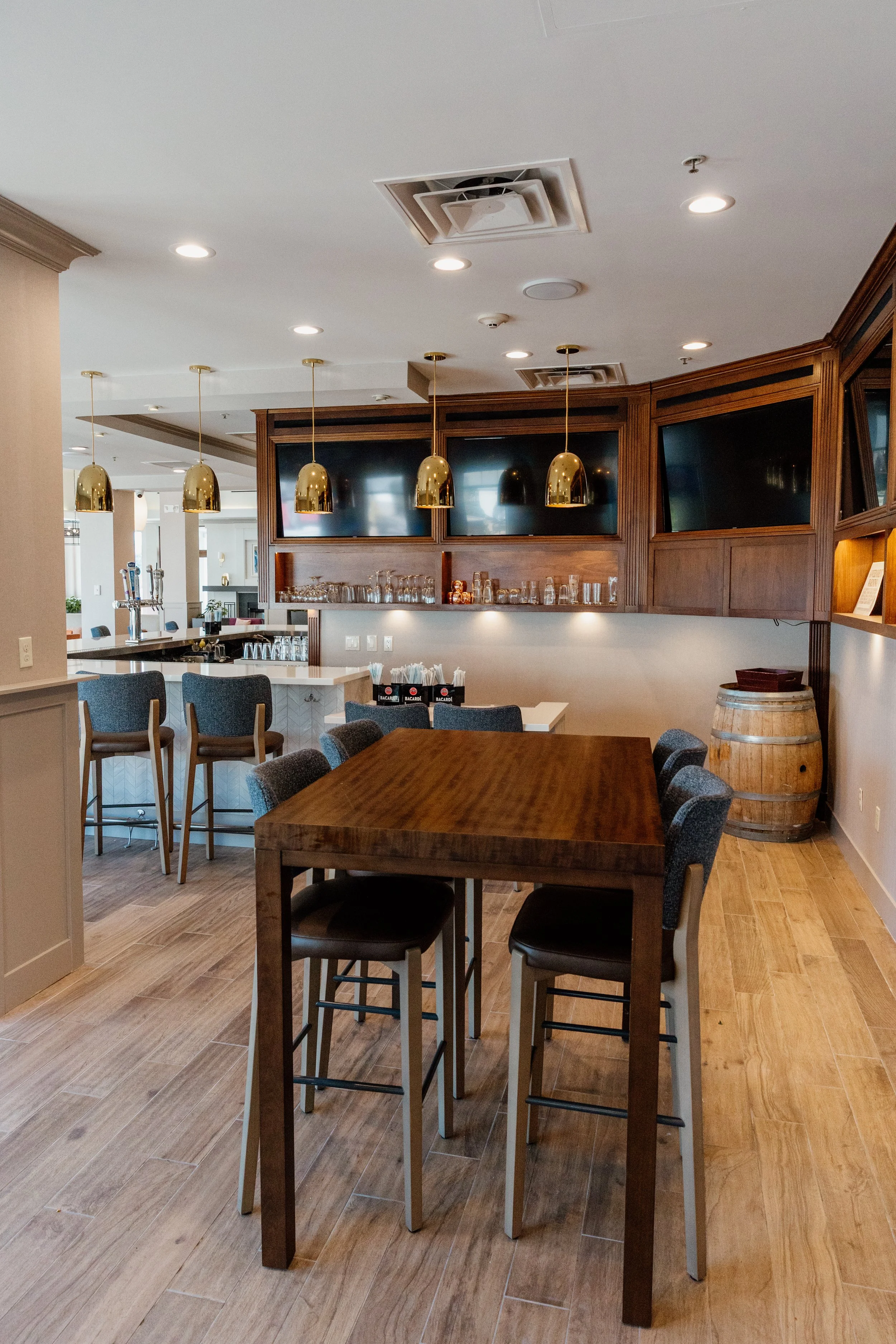 Interior view of a bar and dining area with a wooden table, high chairs, a bar counter with barstools, and a wine barrel. Overhead pendant lights and wall-mounted TVs are visible.