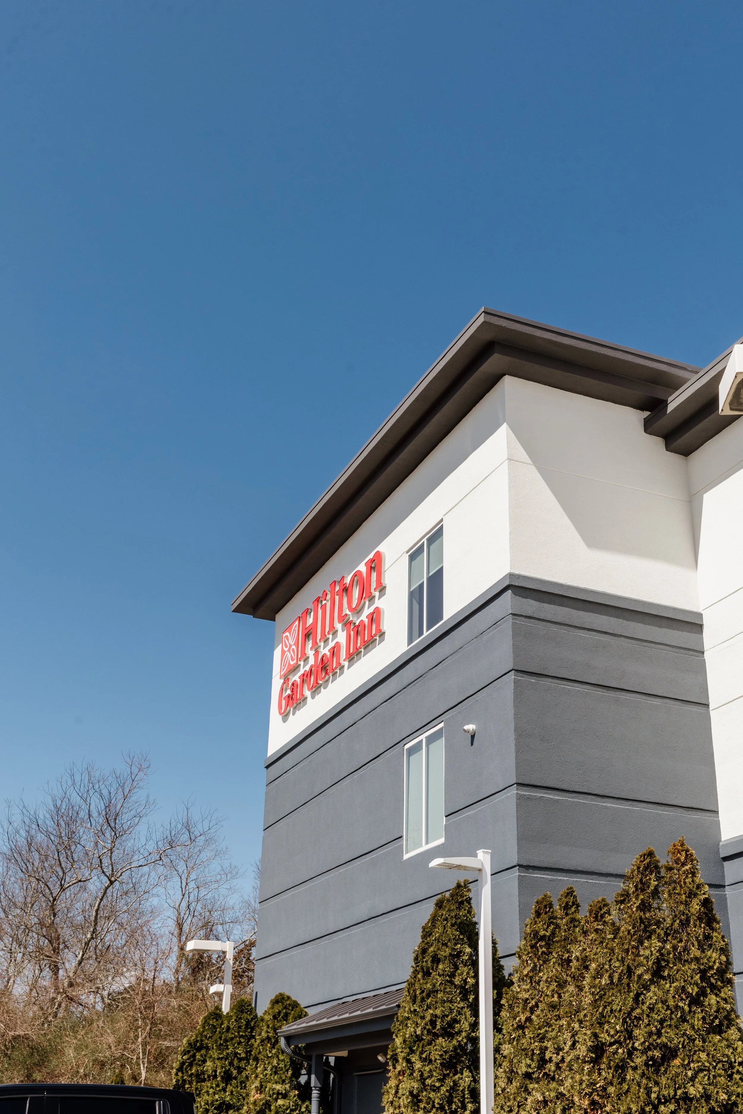 Exterior of a Hilton Garden Inn hotel building with the sign visible, trees, and a clear blue sky.