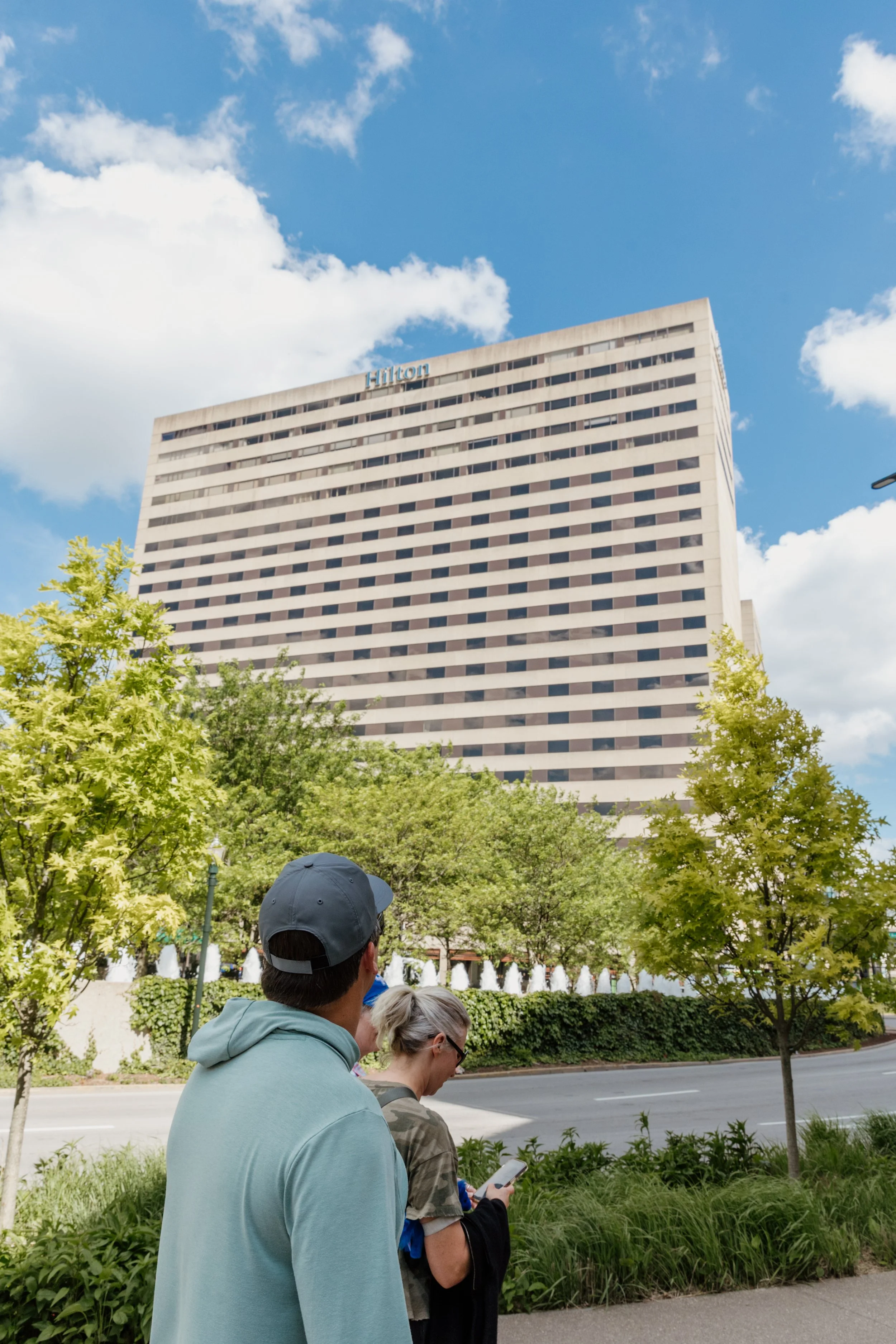 People walking on a sidewalk in front of a large, white office building with the Hilton logo, under a partly cloudy sky, with green trees and bushes along the street.