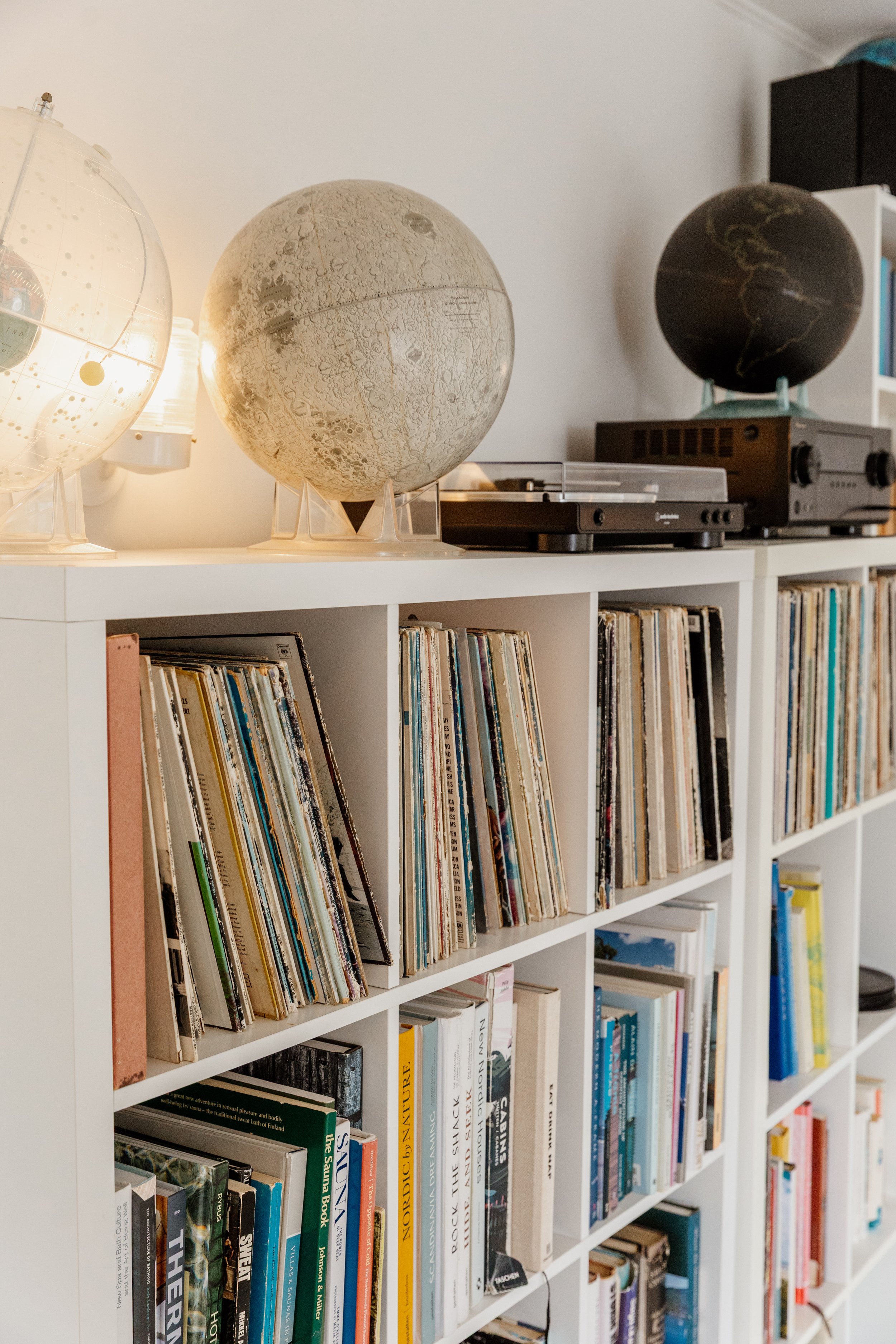 Bookshelf with vinyl records on top, including a moon lamp and globe lamps, and a turntable in a room with white walls.
