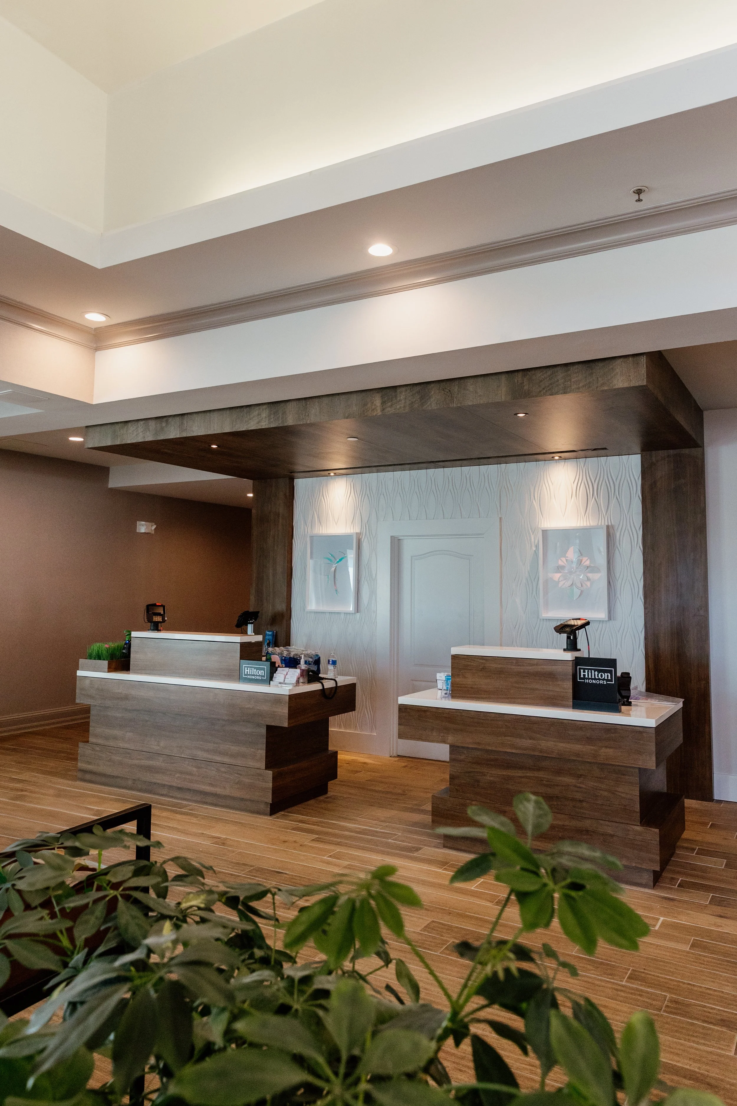 The check-in area at the Hilton Garden Inn in Riverhead, New York, with warm wooden reception counters, framed artwork on the wall, and lush green plants adding a welcoming touch.