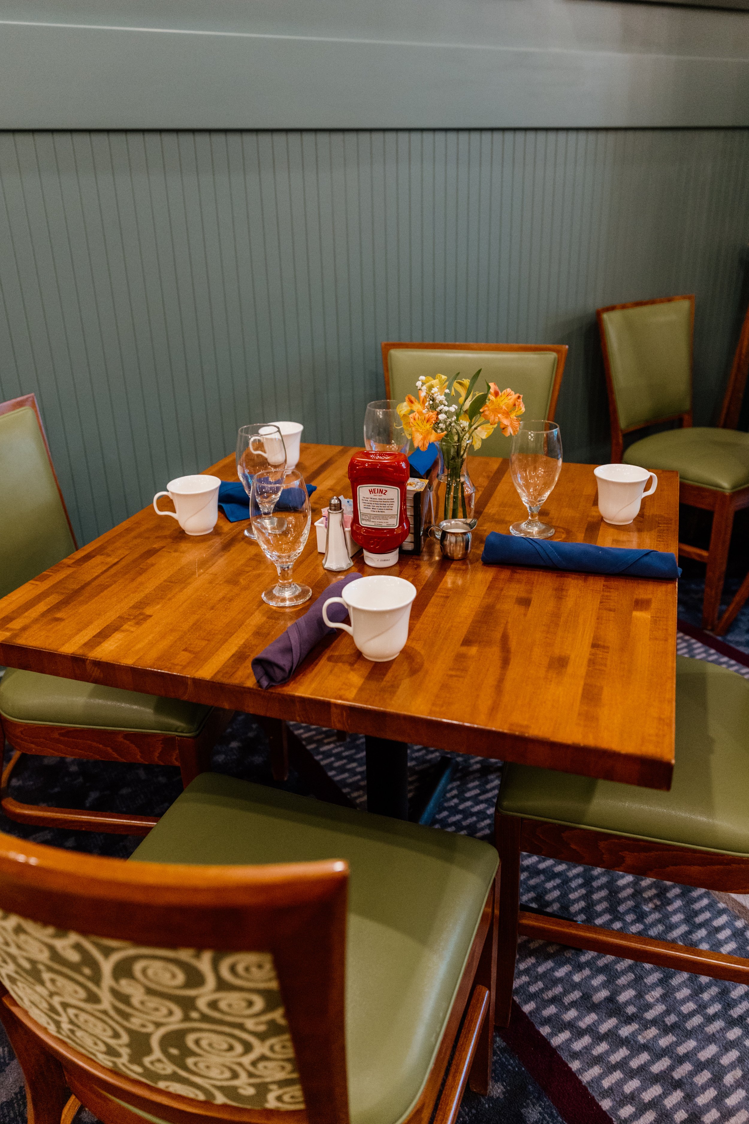 Empty restaurant table with green cushioned chairs, a vase of flowers, four coffee mugs, three wine glasses, a ketchup bottle, salt and pepper shakers, and folded blue and purple napkins.