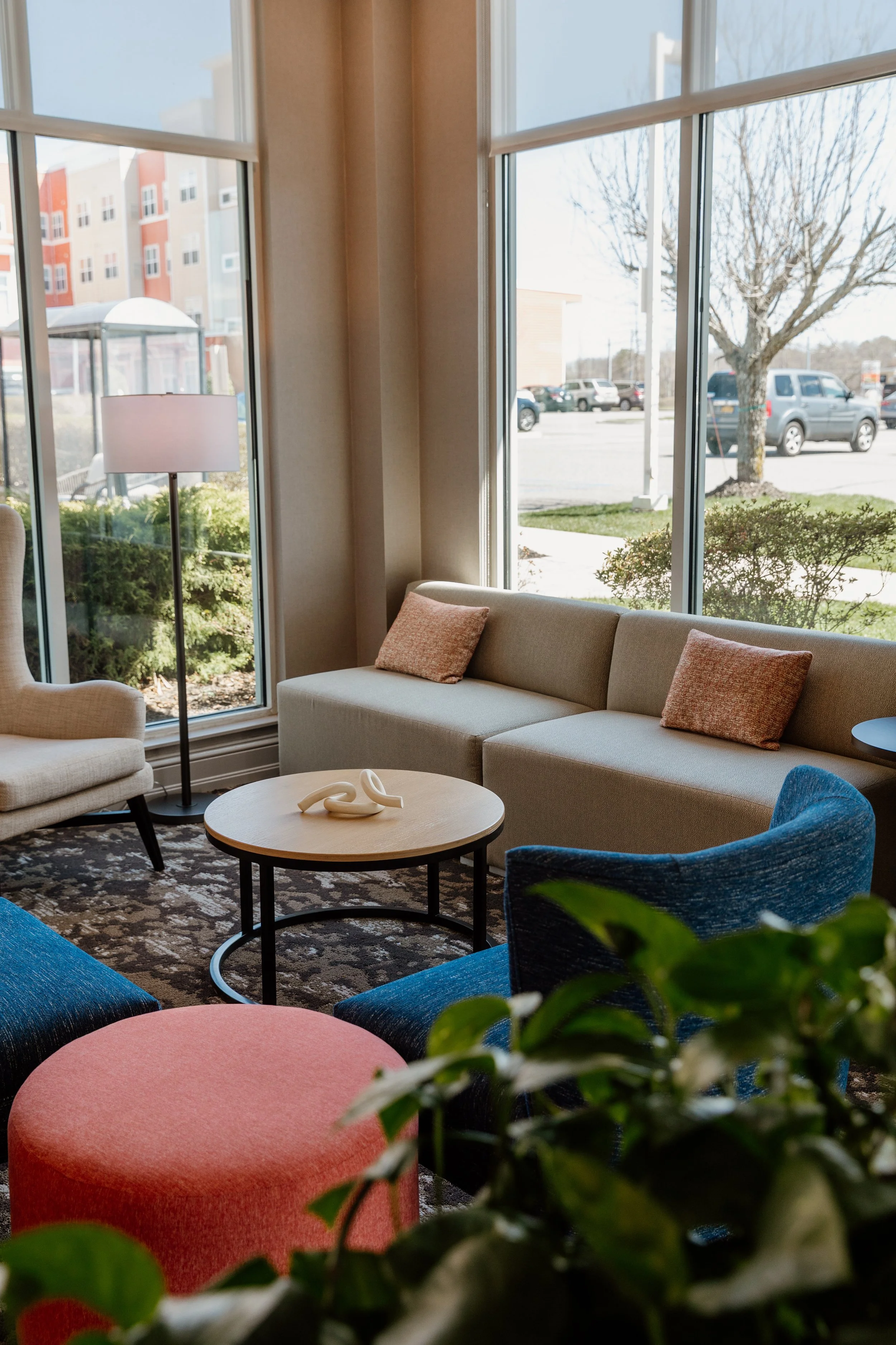 Indoor seating area with a beige sectional sofa, a white armchair, colorful chairs, a round wooden coffee table, a floor lamp, and large windows overlooking outside parking lot and tree.