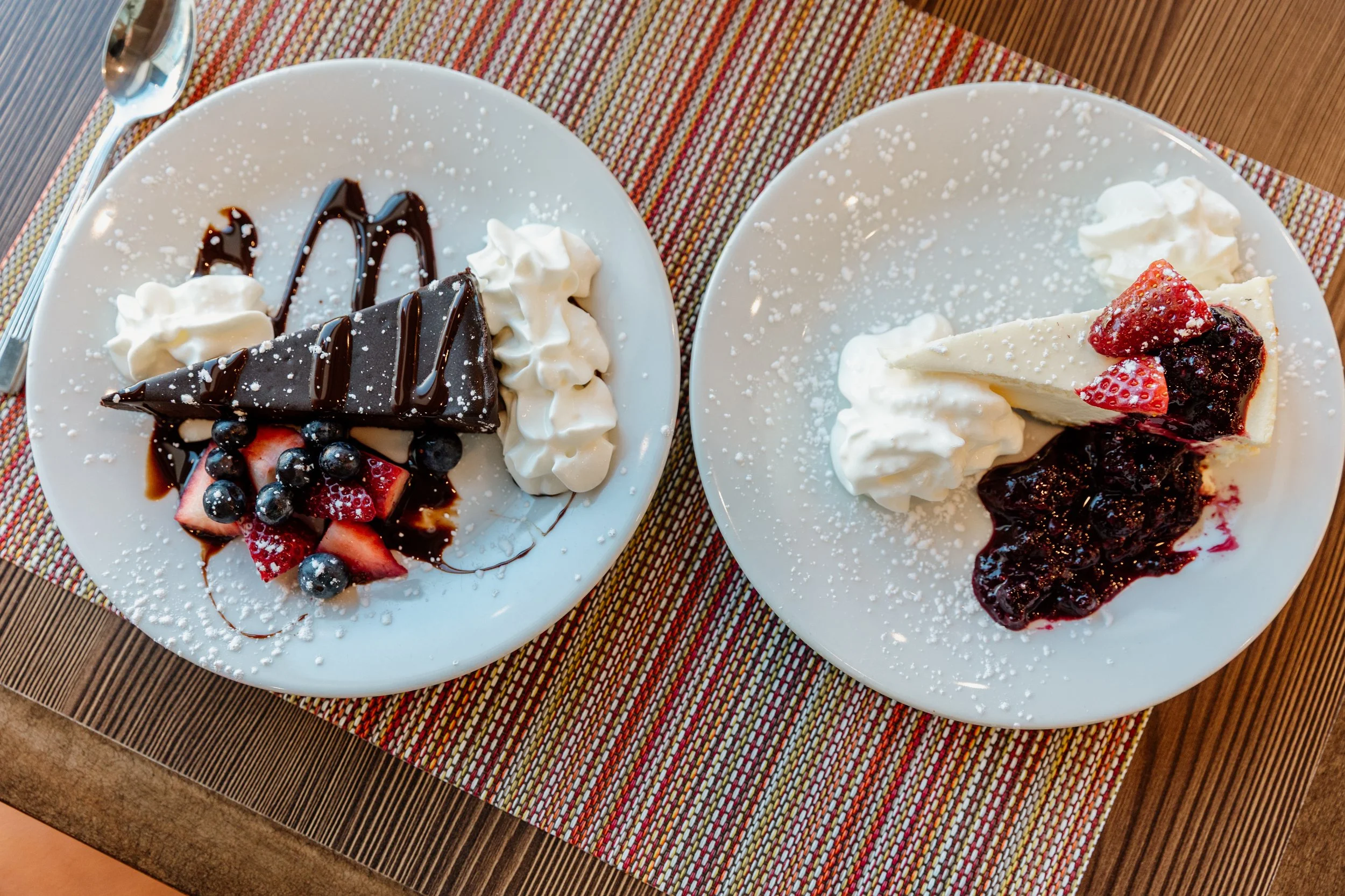 Two plates of cheesecake topped with whipped cream and berries, one with chocolate and the other with fruit preserves.