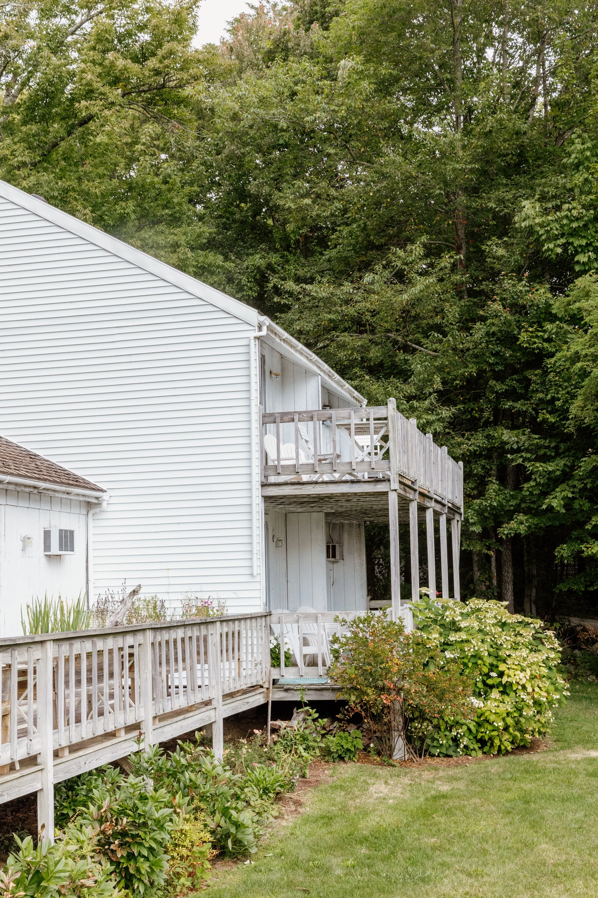 A white two-story house with a wooden balcony and porch, surrounded by green bushes and trees in the backyard.