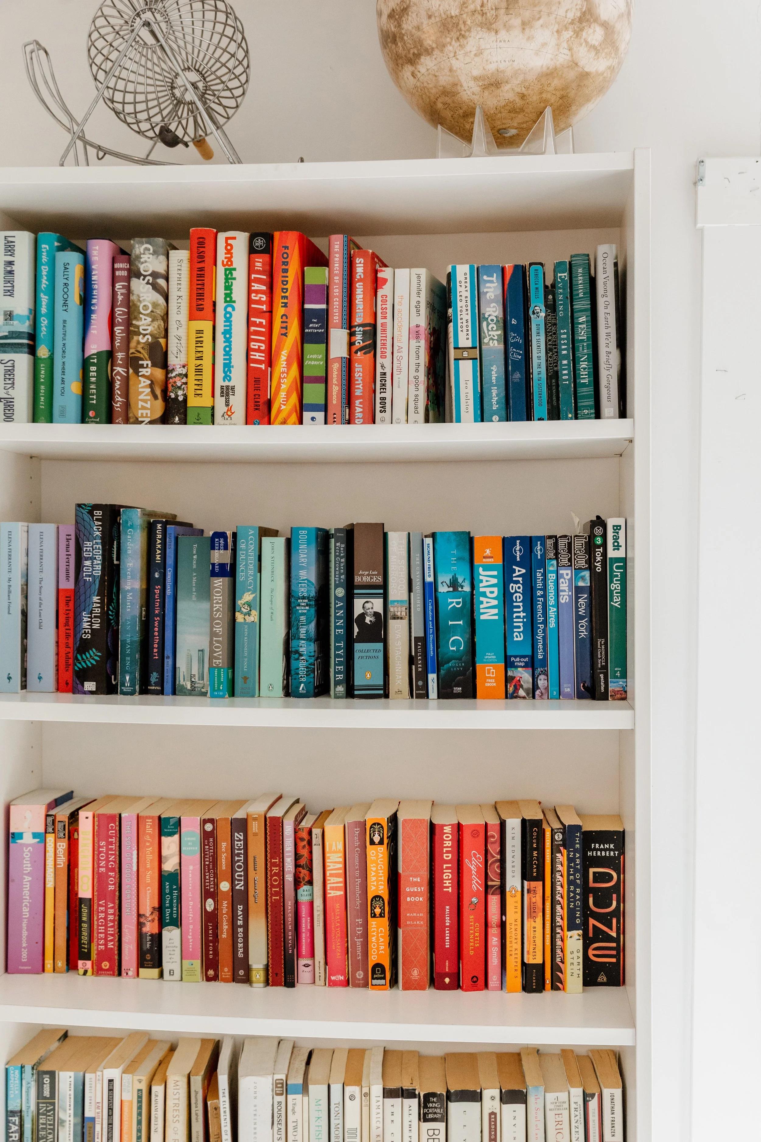 A white bookshelf filled with various colored books. On top of the bookshelf are decorative items including a globe and two vintage fans.