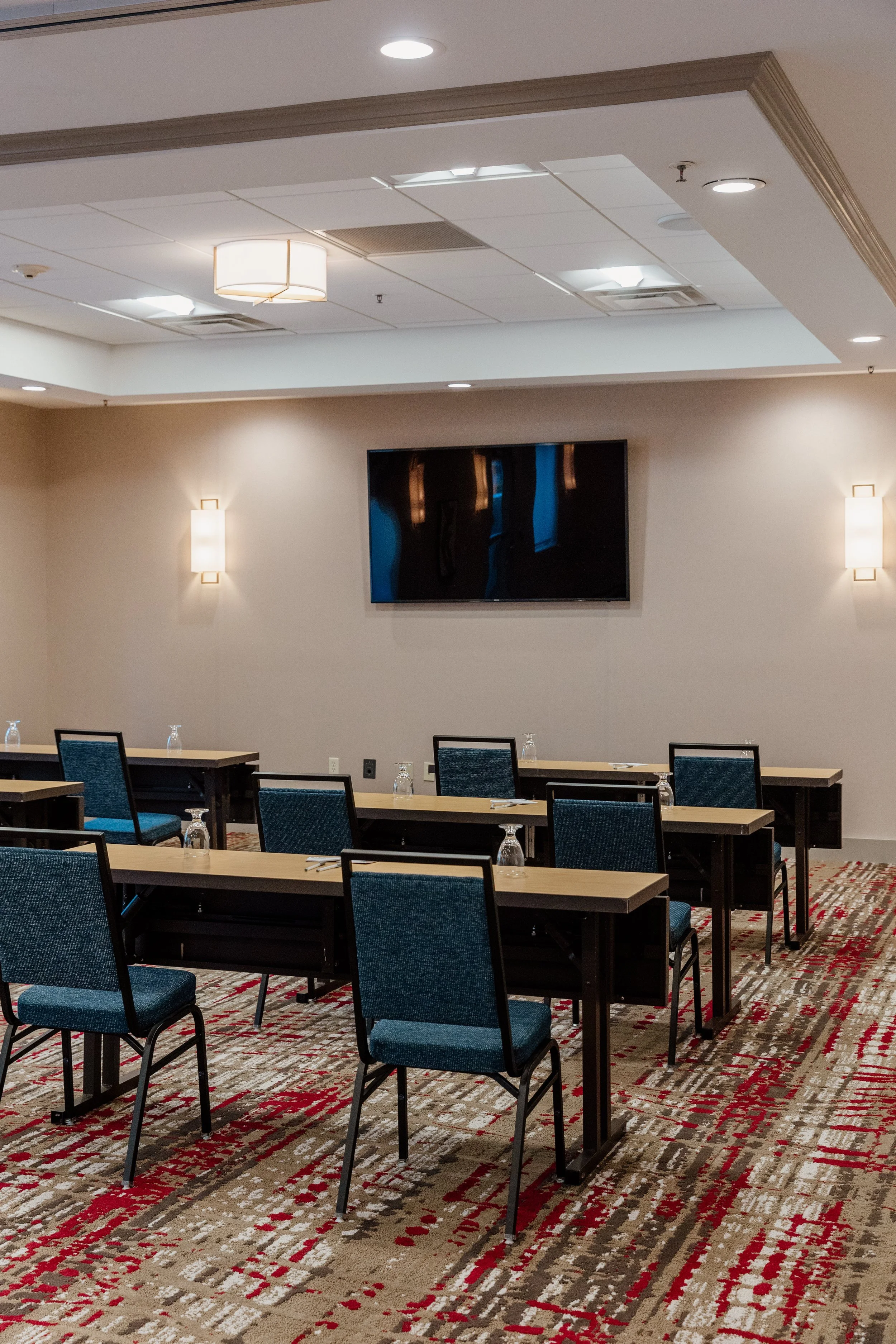 An empty conference room with blue chairs and a long table set with water glasses, facing a wall-mounted flat screen TV, with beige walls and a multi-colored rug.