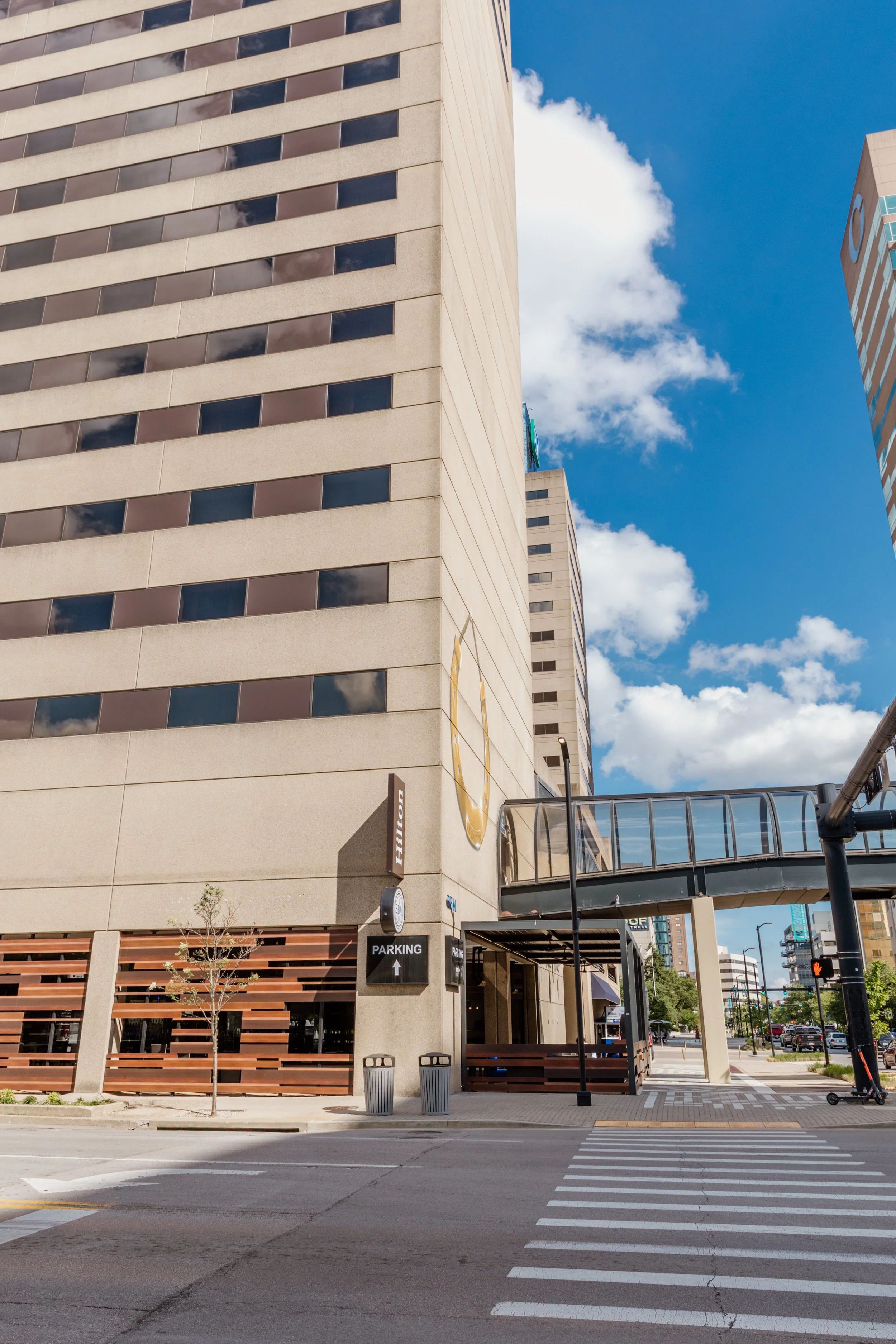 Tall beige and brown office building with reflections of the cloudy blue sky in the windows. A pedestrian bridge connects the building to another structure. A stoplight, trees, and pedestrian crosswalk are visible at the street corner.
